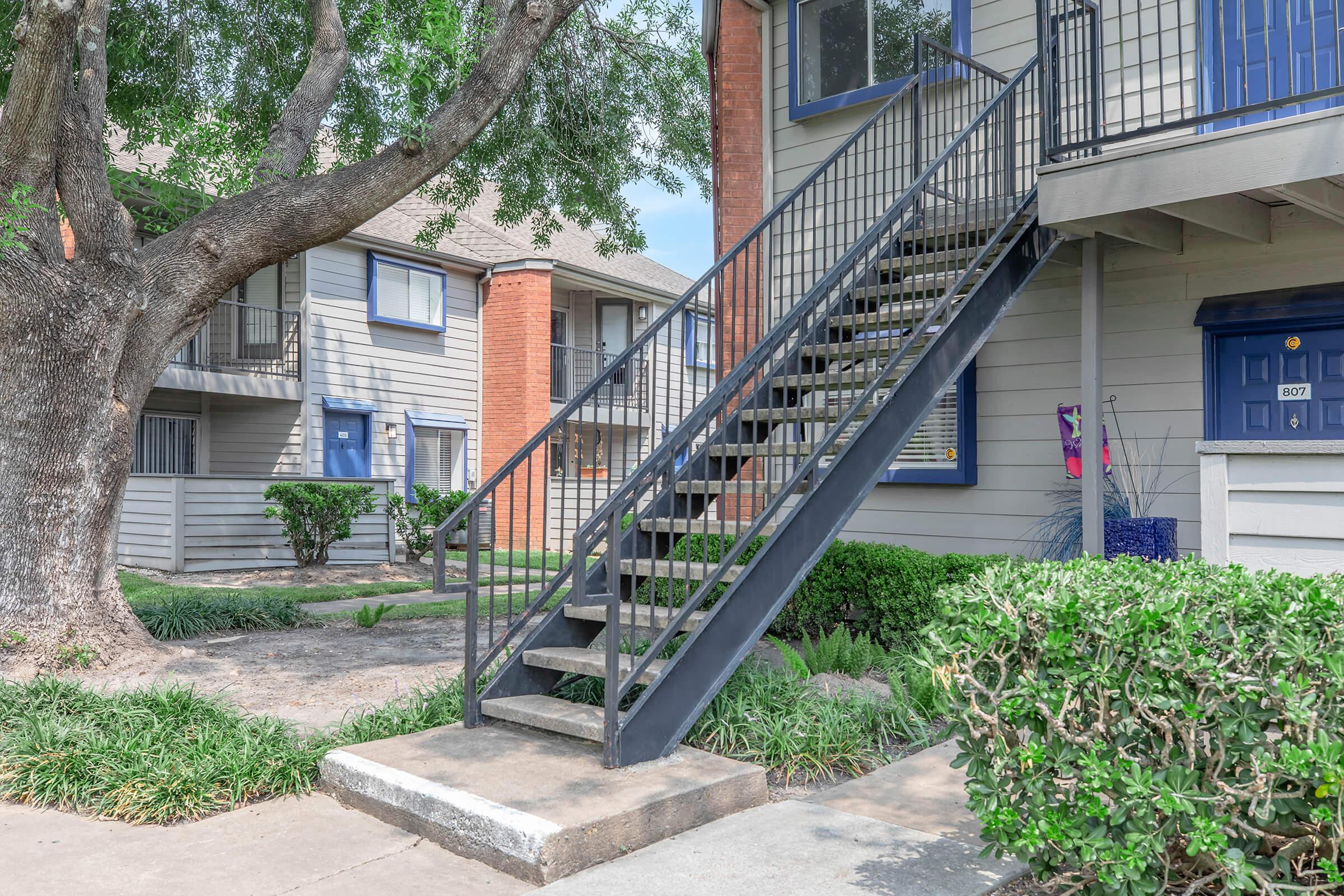Staircase leading to the entrance of an apartment building, surrounded by greenery, with a tree nearby. Two attached apartment units are visible in the background, featuring blue doors and windows. The setting is well-maintained and reflects a residential area.
