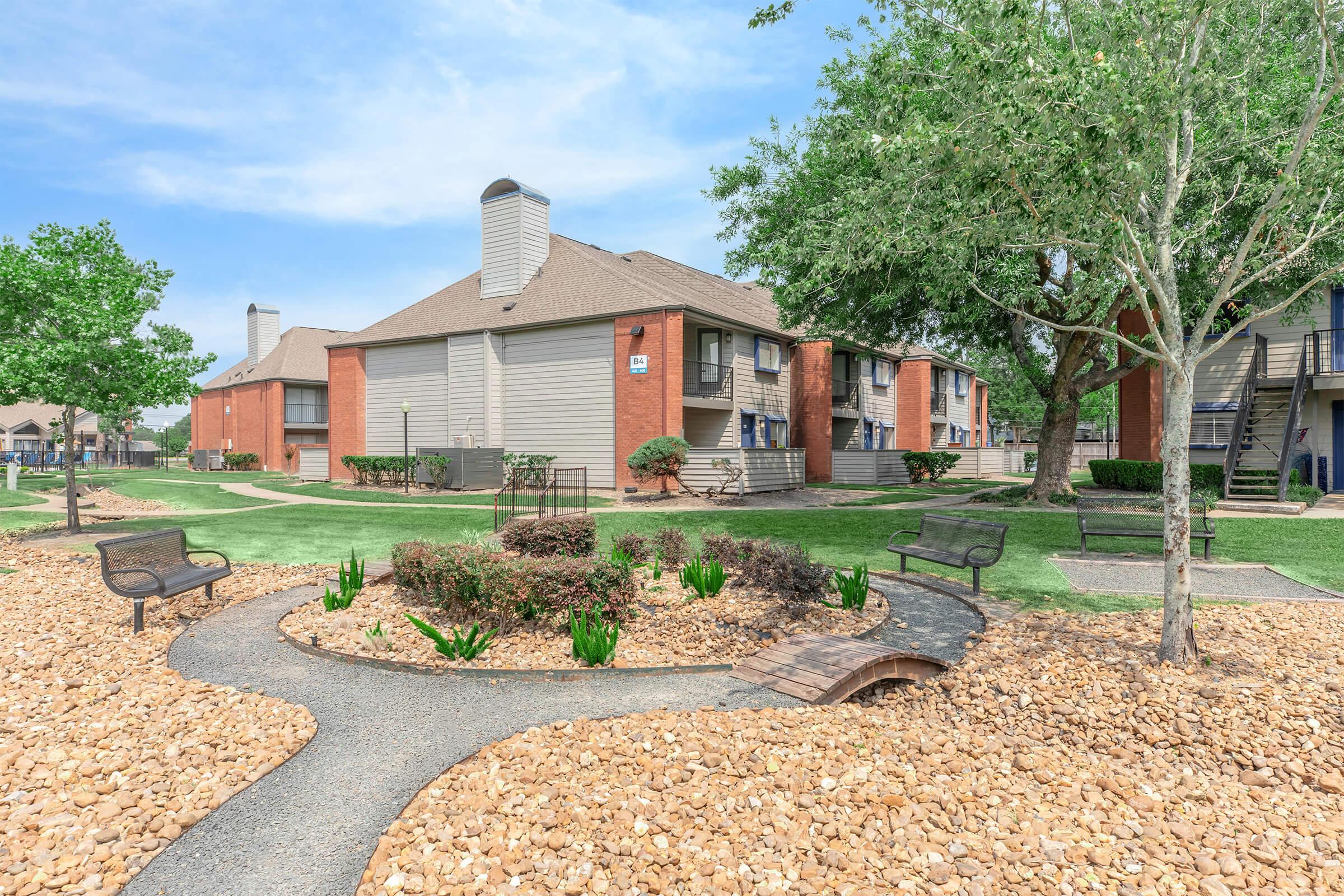 A landscaped area featuring a pathway made of gravel that winds through garden beds with shrubs and flowers. In the background, there are brown multi-unit apartment buildings with sloped roofs. Green grass and trees enhance the outdoor setting, creating a serene residential atmosphere.
