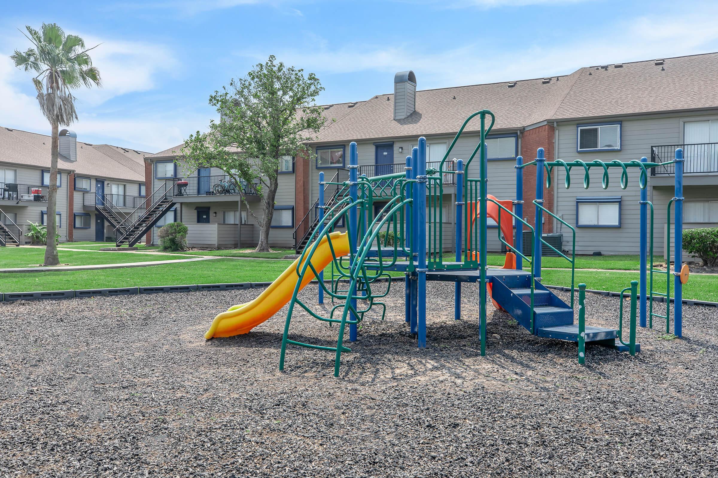 Playground equipment featuring a bright yellow slide and green climbing structures, set in a landscaped area with grass and palm trees. In the background, there are multi-story residential buildings with balconies. The scene is well-lit and inviting for children.