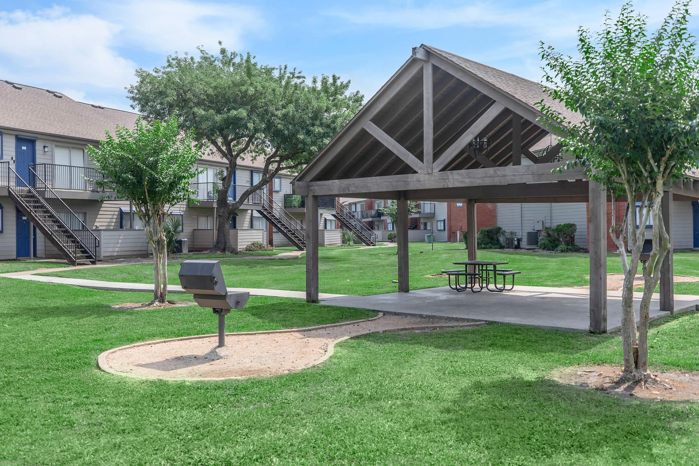 A grassy courtyard featuring a large shaded pavilion with picnic tables, surrounded by trees and two-story apartment buildings. A charcoal grill is situated nearby, making it a suitable space for outdoor gatherings and relaxation. Clear blue skies enhance the inviting atmosphere.