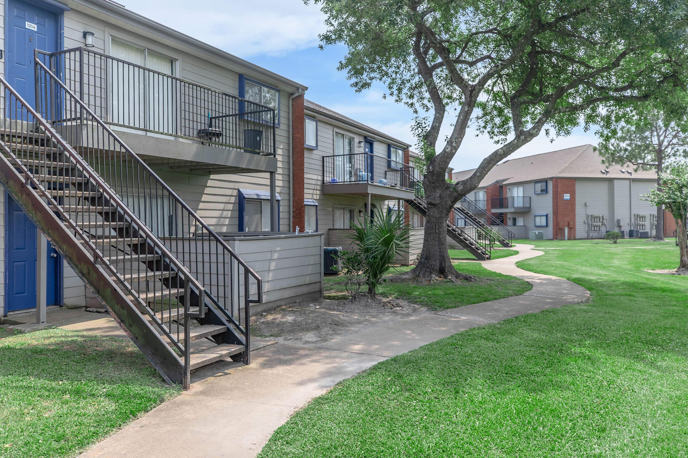 Two two-story apartment buildings with exterior stairs and balconies, surrounded by a grassy area. A winding concrete pathway leads through the landscape, with a tree and some shrubs nearby. The buildings have a mix of colors, with one side painted in shades of beige and another in red.