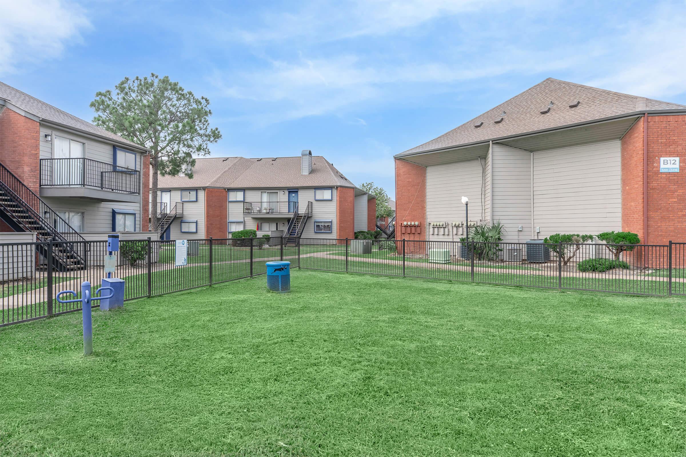 View of a residential apartment complex featuring two buildings with brick exteriors. The foreground includes a grassy area and a blue trash bin. Black railings and staircases are visible, alongside neatly trimmed hedges, creating a well-maintained outdoor space.