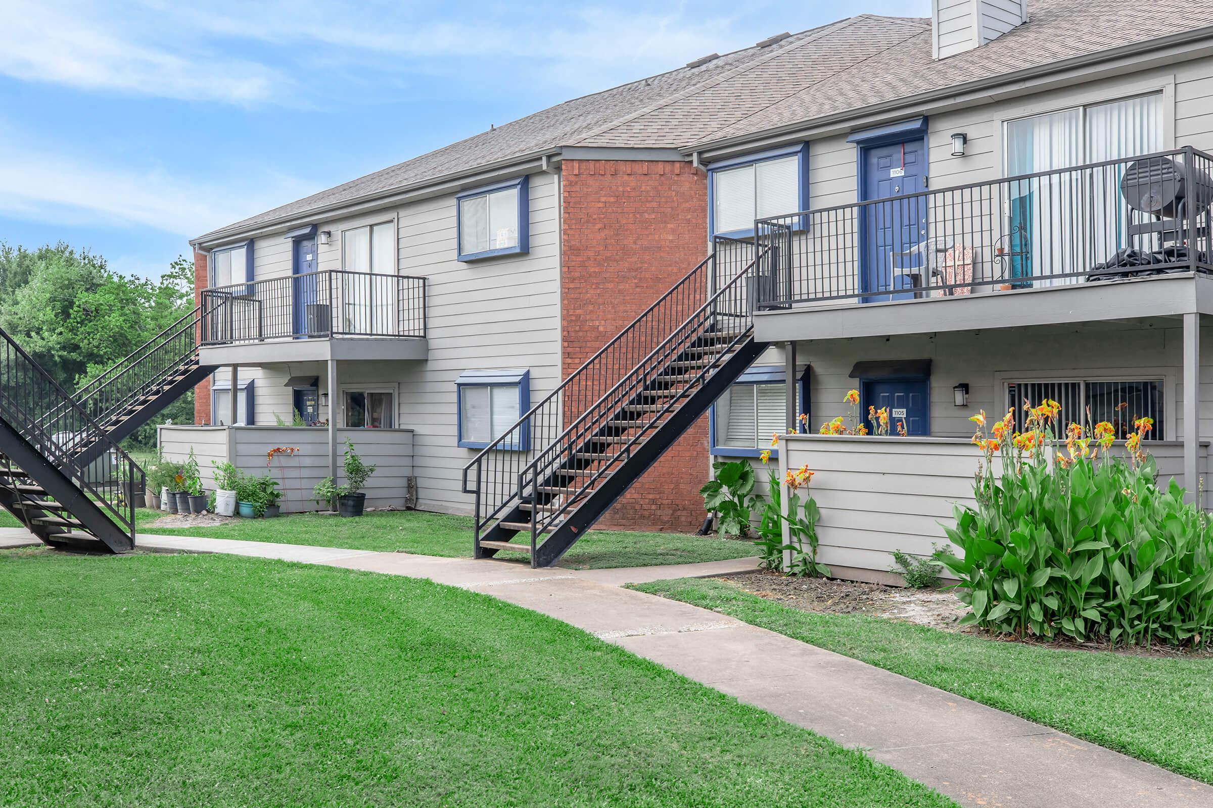 Two-story apartment building with exterior stairs leading to entrance doors. Each unit features a small balcony and landscaped area with blooming flowers. A well-maintained lawn and walkway enhance the inviting appearance of the complex under a clear blue sky.
