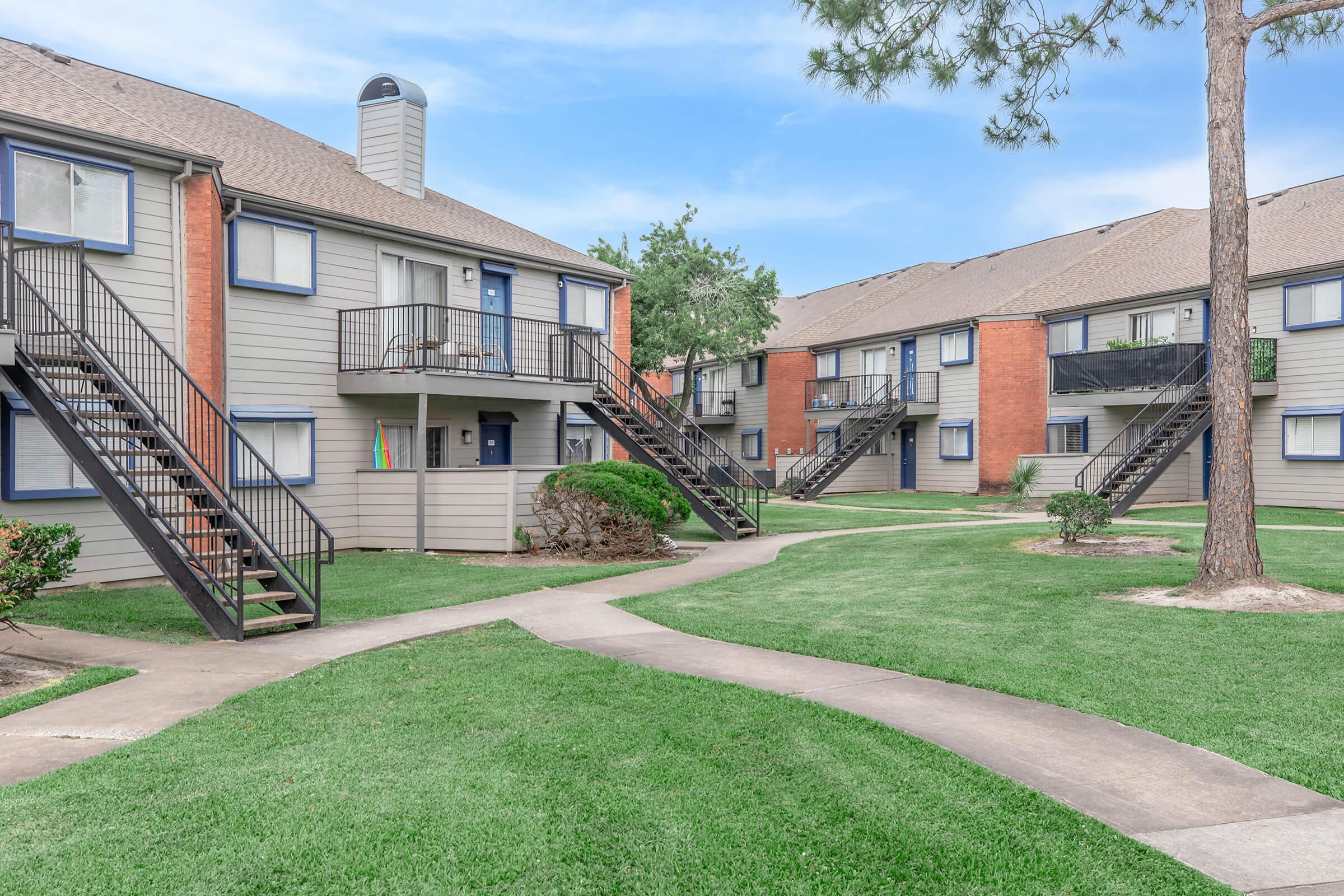 Two-story apartment buildings with gray siding and brick accents, featuring balconies and staircases. Well-maintained green lawns surround the buildings, with a walkway leading through the area. The sky is clear, and a few trees are visible, adding to the pleasant outdoor atmosphere.
