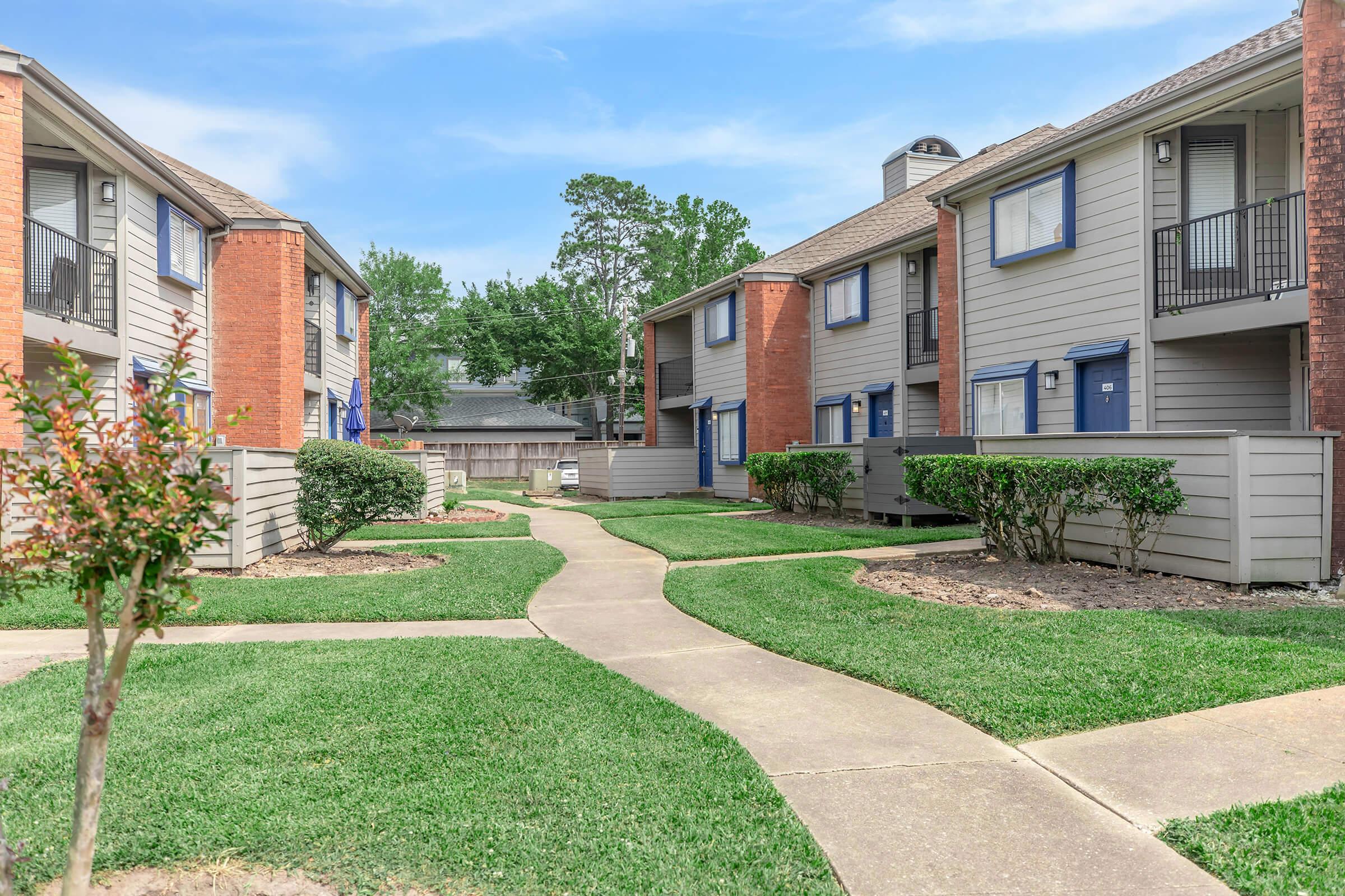 A view of a well-maintained apartment complex featuring two-story buildings with gray siding and red brick accents. Lush green grass borders winding pathways that connect the units, surrounded by neatly trimmed shrubs. Clear blue skies enhance the inviting atmosphere of the outdoor space.