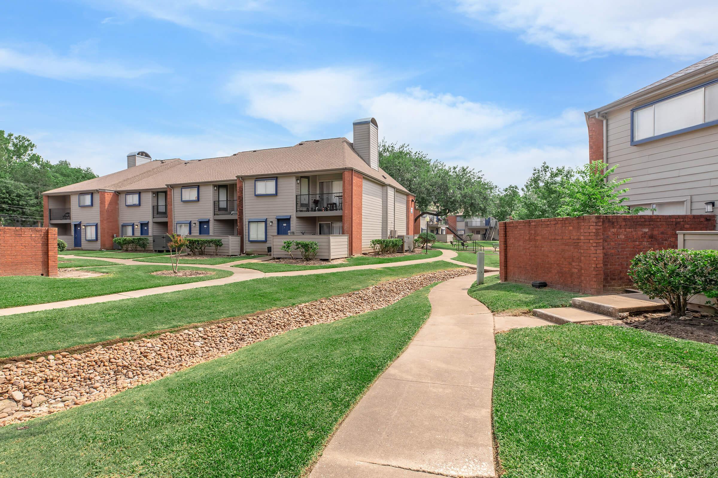 A well-maintained residential complex featuring two multi-story buildings with balconies, surrounded by neatly manicured lawns and pathways. The landscape includes trees and shrubbery, creating a peaceful outdoor environment. Bright blue sky overhead enhances the inviting atmosphere.
