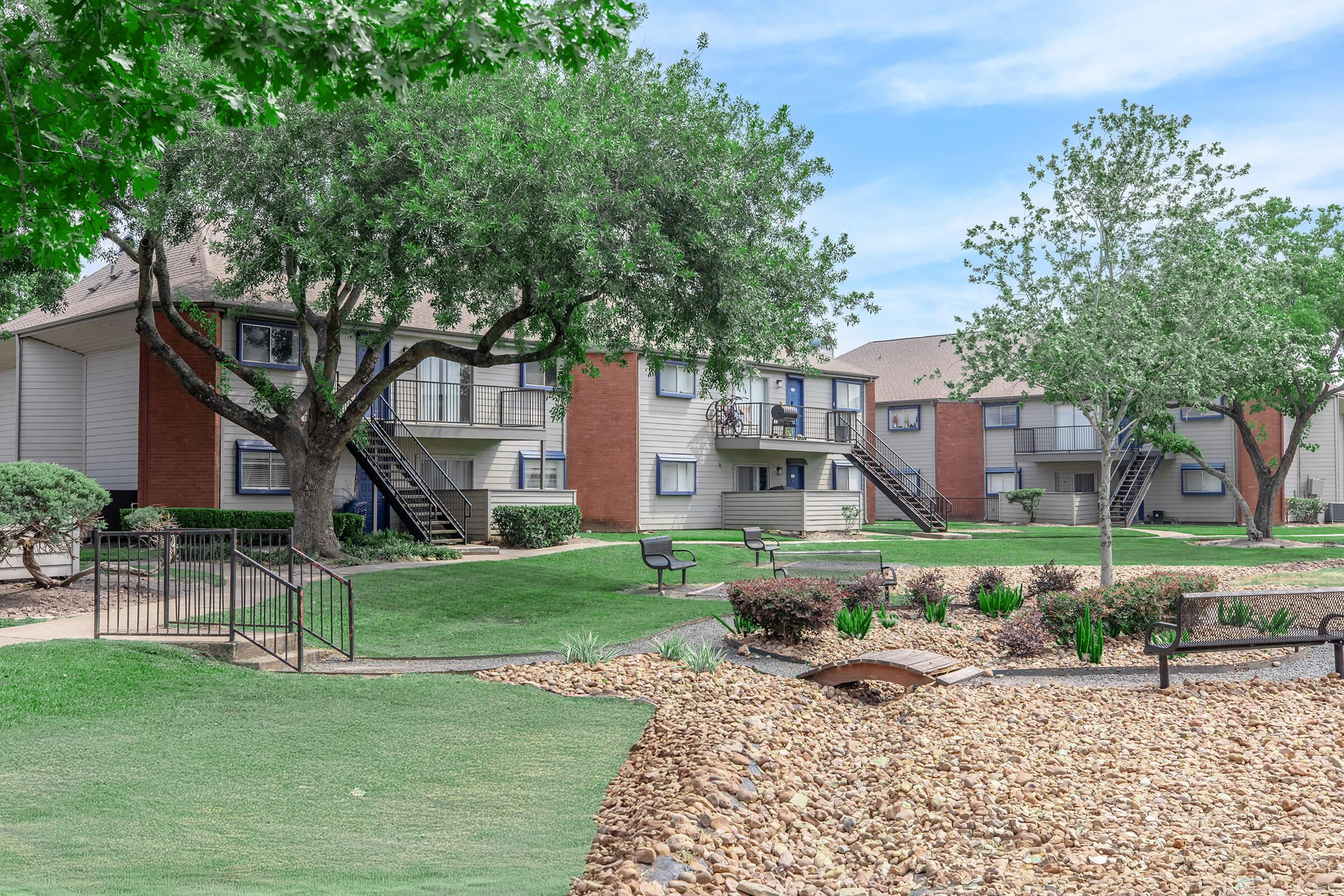 A landscaped residential complex featuring two-story apartment buildings with red brick and light-colored siding. The area includes lush green lawns, trees, and flower beds, with benches and pathways visible. A clear blue sky is seen in the background, creating a serene atmosphere.