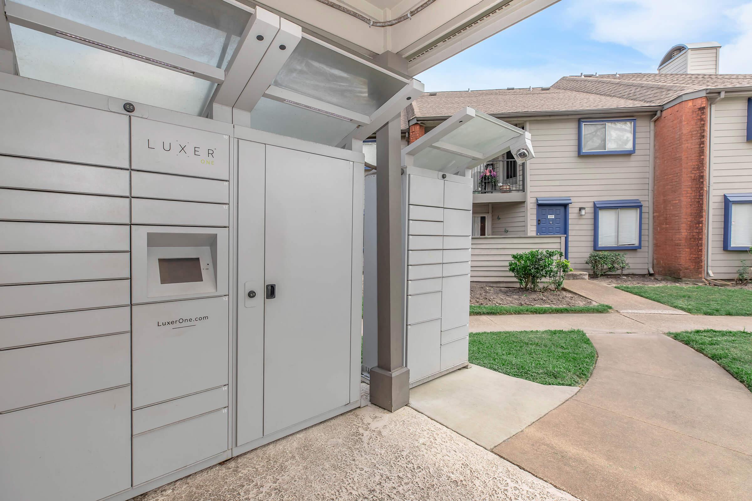 Package delivery lockers situated outside an apartment building. The lockers feature a sleek design with a digital keypad. Lush green grass surrounds the area, and there are walkways leading to nearby apartment entrances, creating a convenient space for residents to pick up their deliveries.