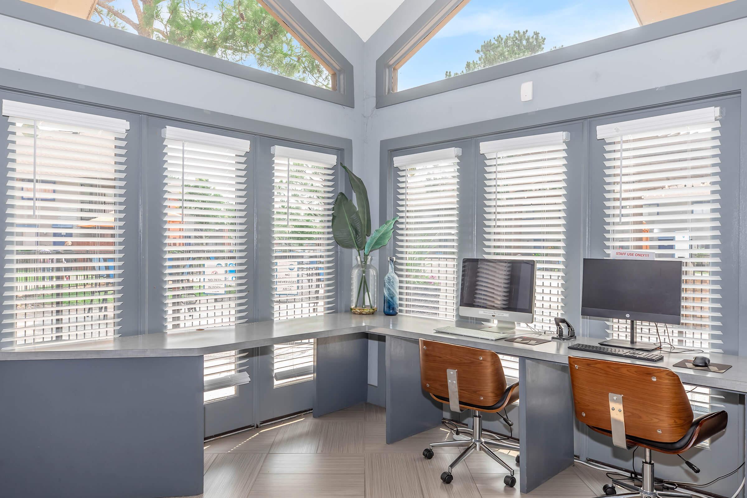 A modern workspace featuring a large corner desk with two computer monitors, chairs, and decorative plants. The room has multiple windows with white blinds, allowing natural light. The walls are painted in a soft gray color, complemented by a stylish geometric floor.