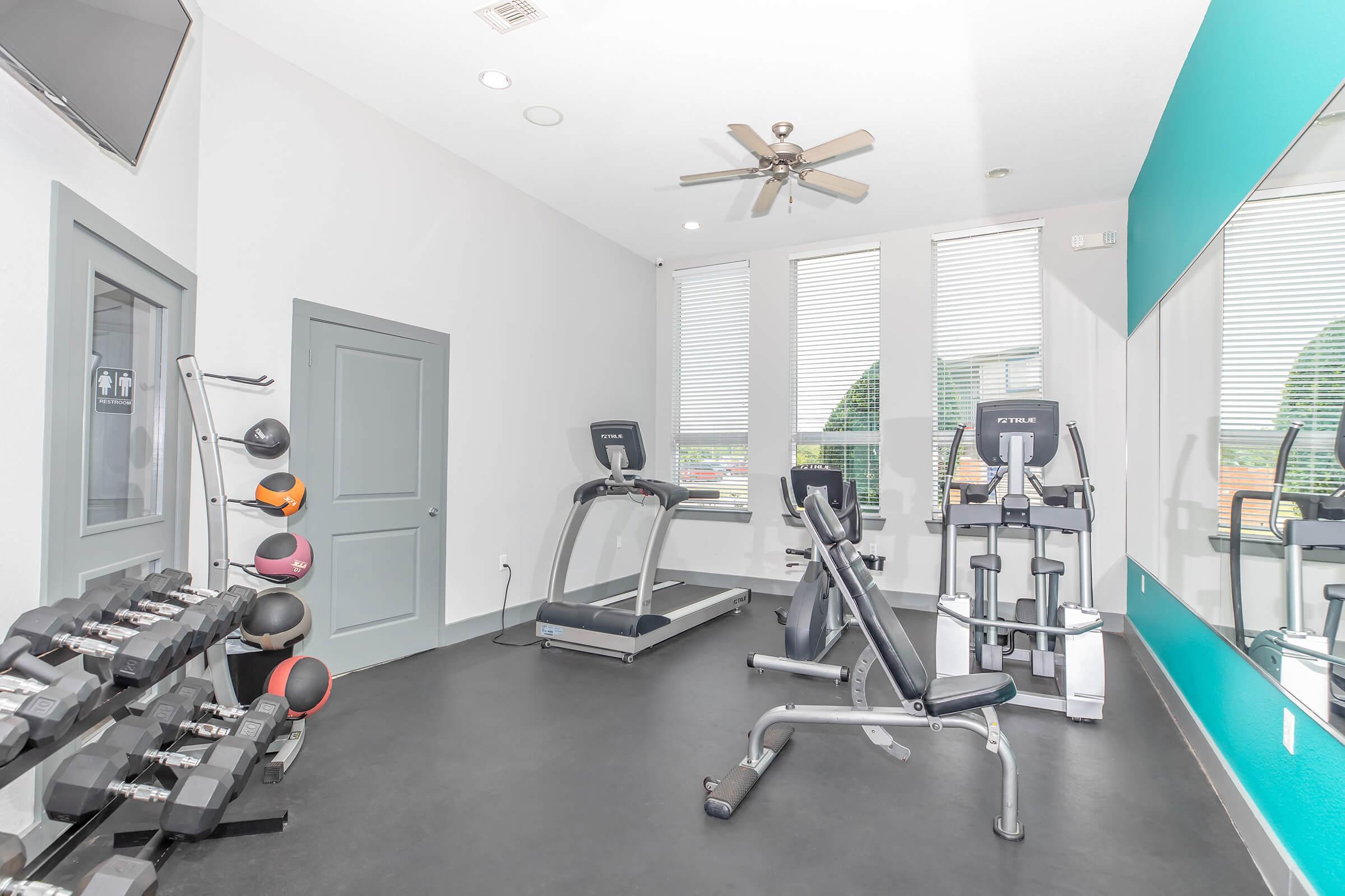 A bright, modern fitness room featuring exercise equipment including treadmills, an elliptical machine, and free weights. Large windows provide natural light, and there are mirrors on one wall, enhancing the spacious feel of the area. A ceiling fan adds comfort to the environment.