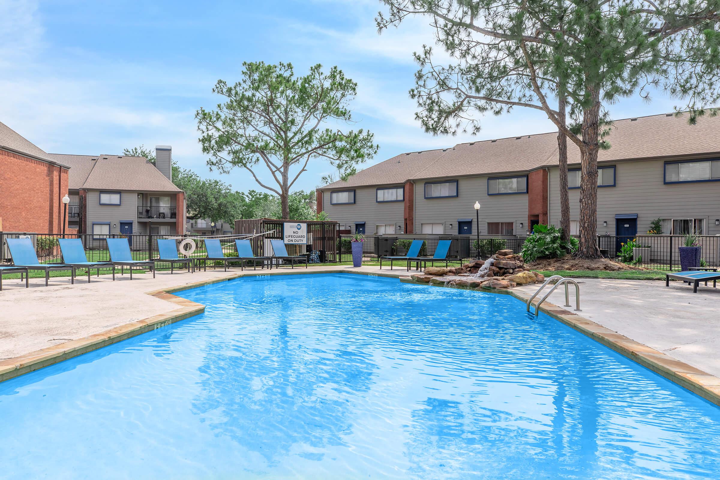 A bright outdoor swimming pool surrounded by lounge chairs, with a few trees and buildings in the background. The water is clear and inviting, reflecting the blue sky. A rock formation adds a natural element near the pool's edge.