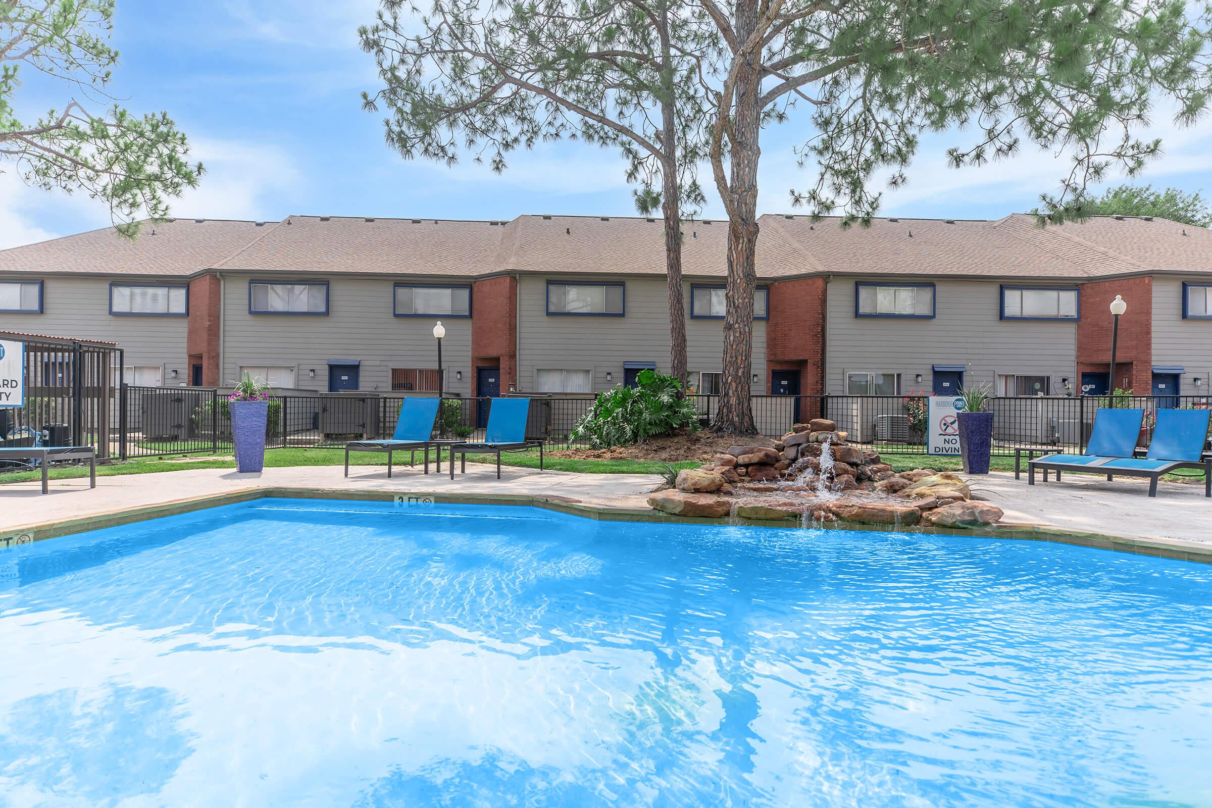 A sparkling blue swimming pool surrounded by lounge chairs and greenery. In the background, there are several two-story townhomes with gray siding and brick accents. A small waterfall feature adds to the tranquil atmosphere, and a few trees provide shade.