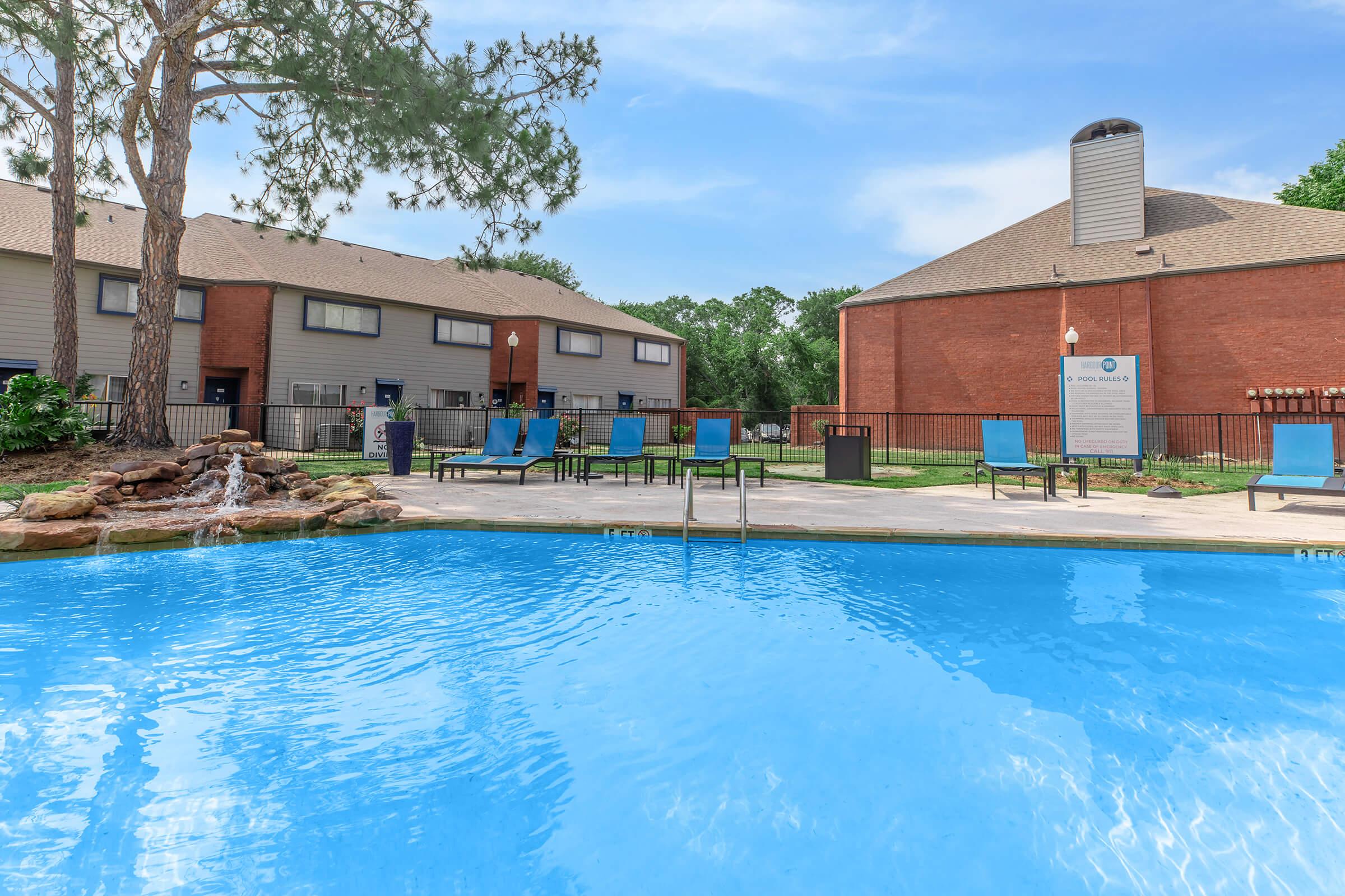 A clear swimming pool with a waterfall feature, surrounded by lounge chairs. In the background, there are two residential buildings with a mix of brick and siding exteriors, and a large tree providing shade. The scene is bright and inviting, showcasing a warm, sunny day.