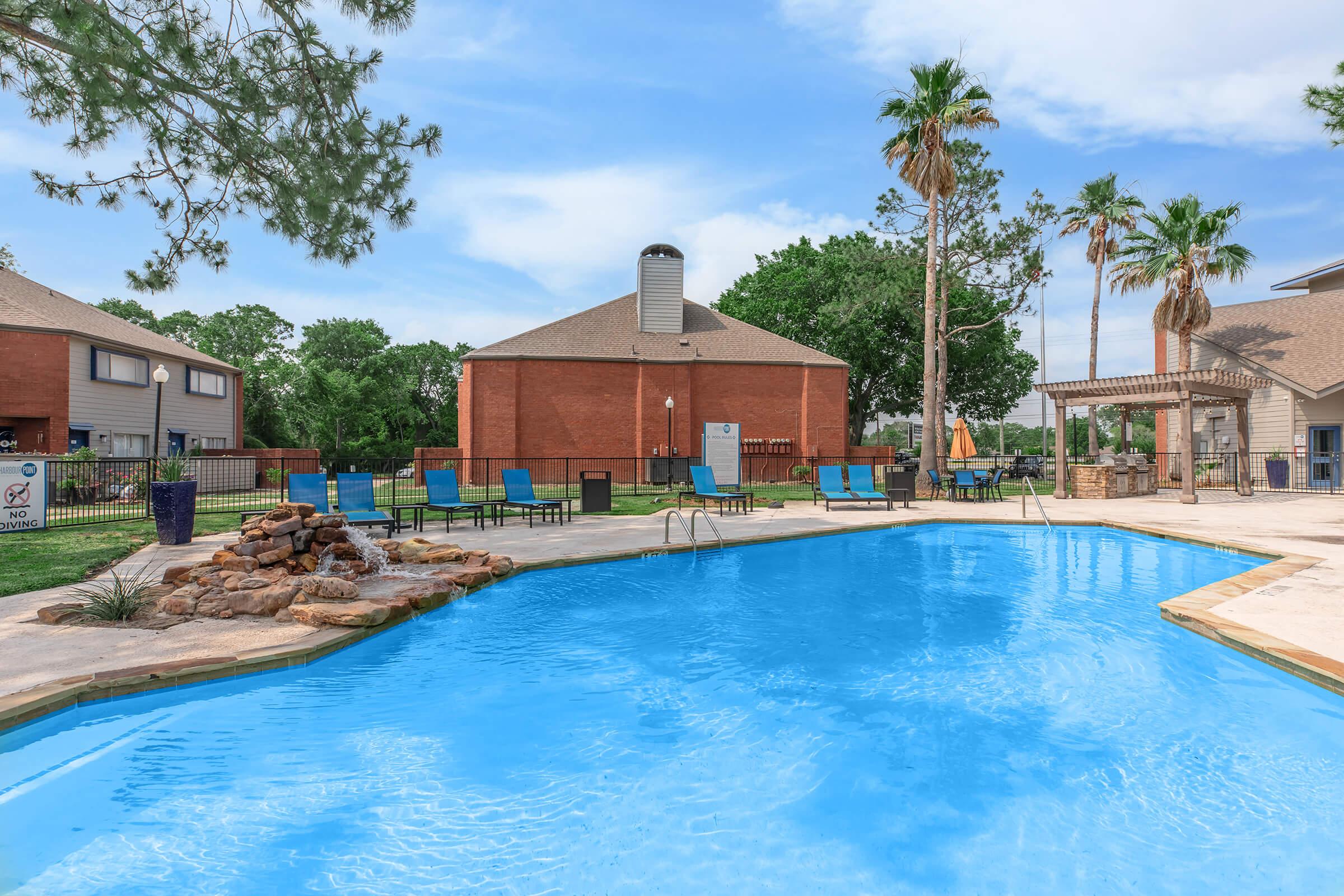 A bright blue swimming pool surrounded by lounge chairs and palm trees, set against a backdrop of brick buildings. There are landscaped areas with rocks near the pool, and the scene is bathed in natural light under a clear sky.