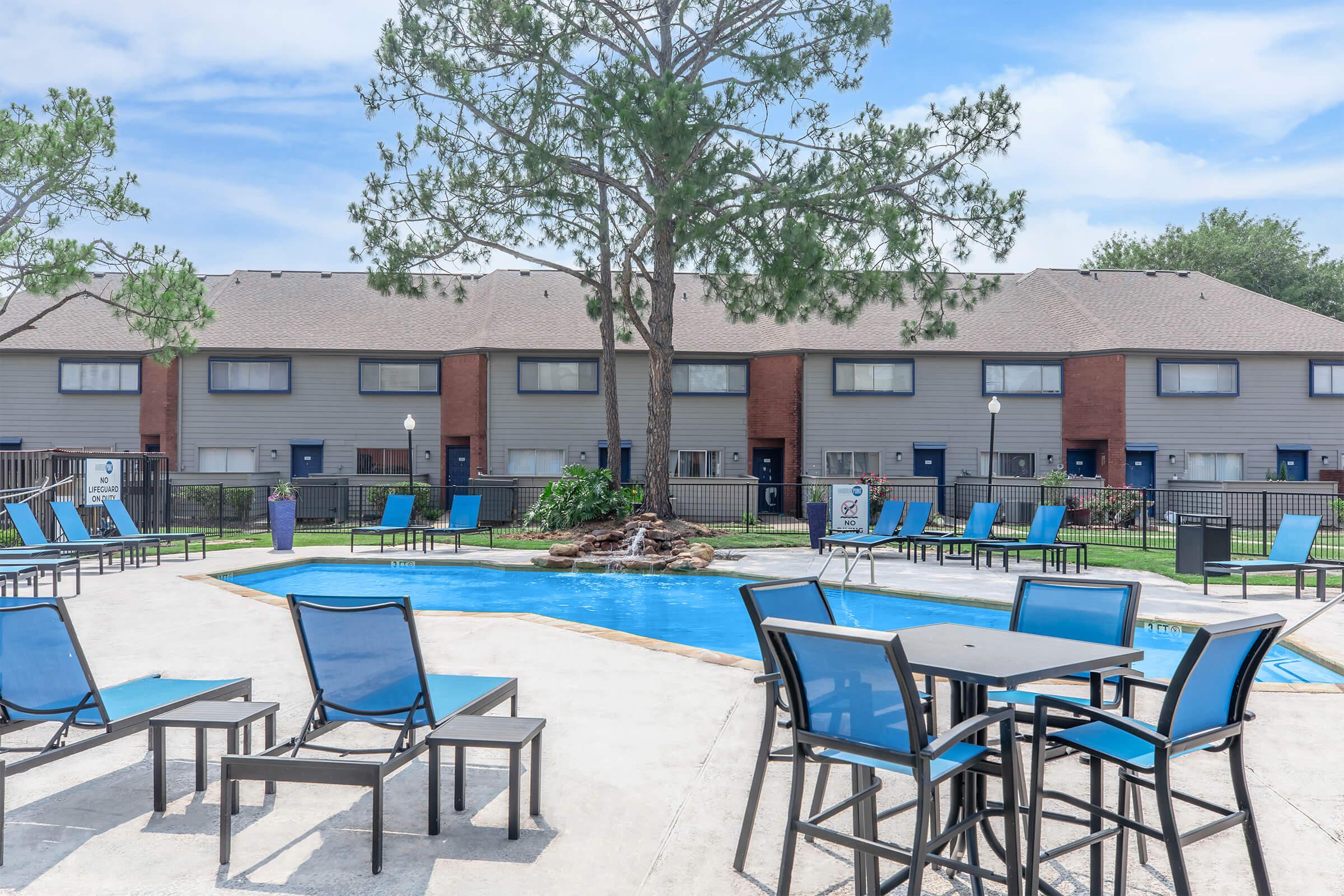 A swimming pool surrounded by lounge chairs and tables in a residential area. The pool features a decorative tree in the center, with buildings visible in the background. Clear blue skies add to the inviting atmosphere of the outdoor space.