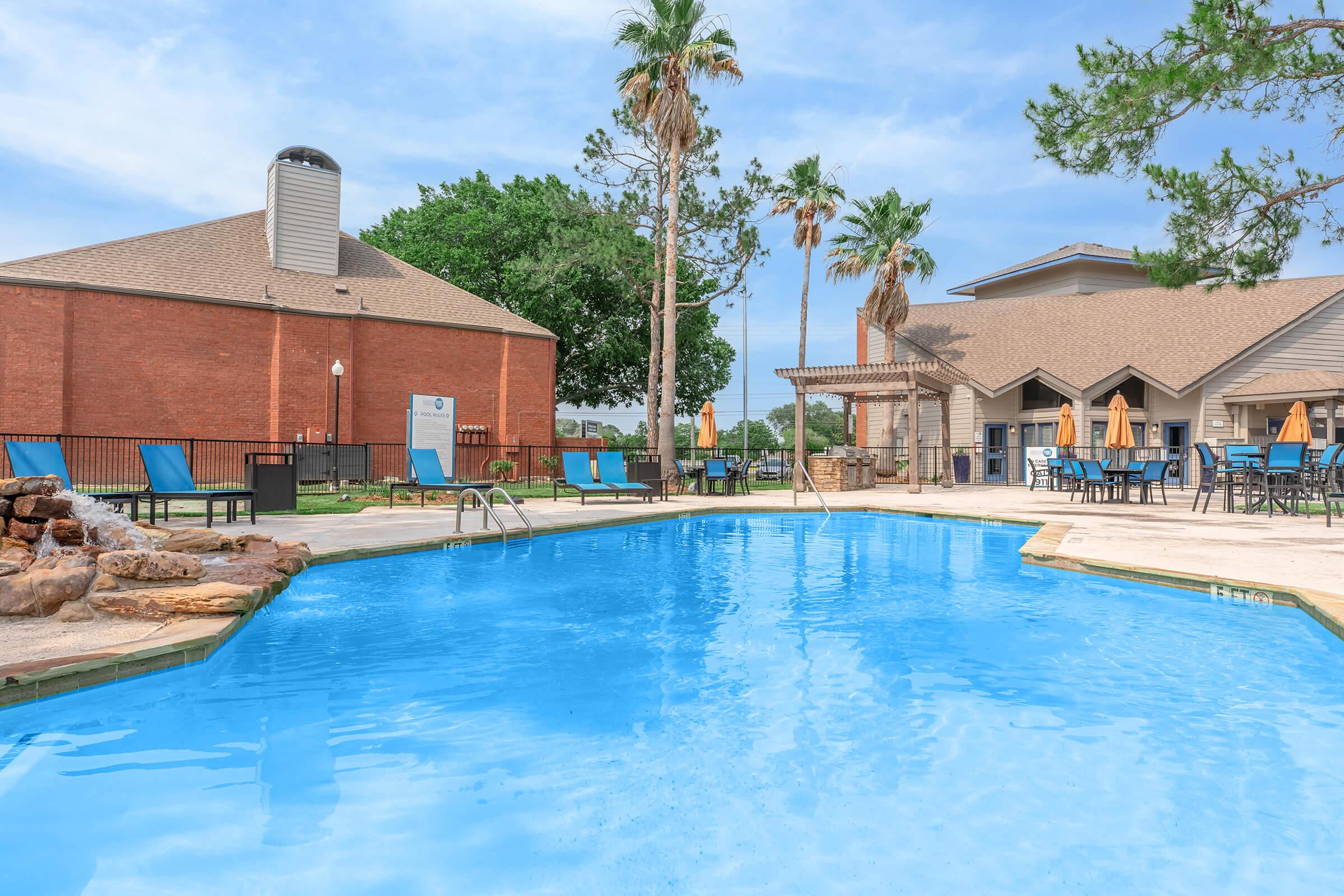 A serene swimming pool surrounded by lounge chairs and palm trees, with a brick building in the background. The pool area features stone accents and multiple umbrellas for shade, creating a relaxing outdoor space ideal for leisure and recreation.