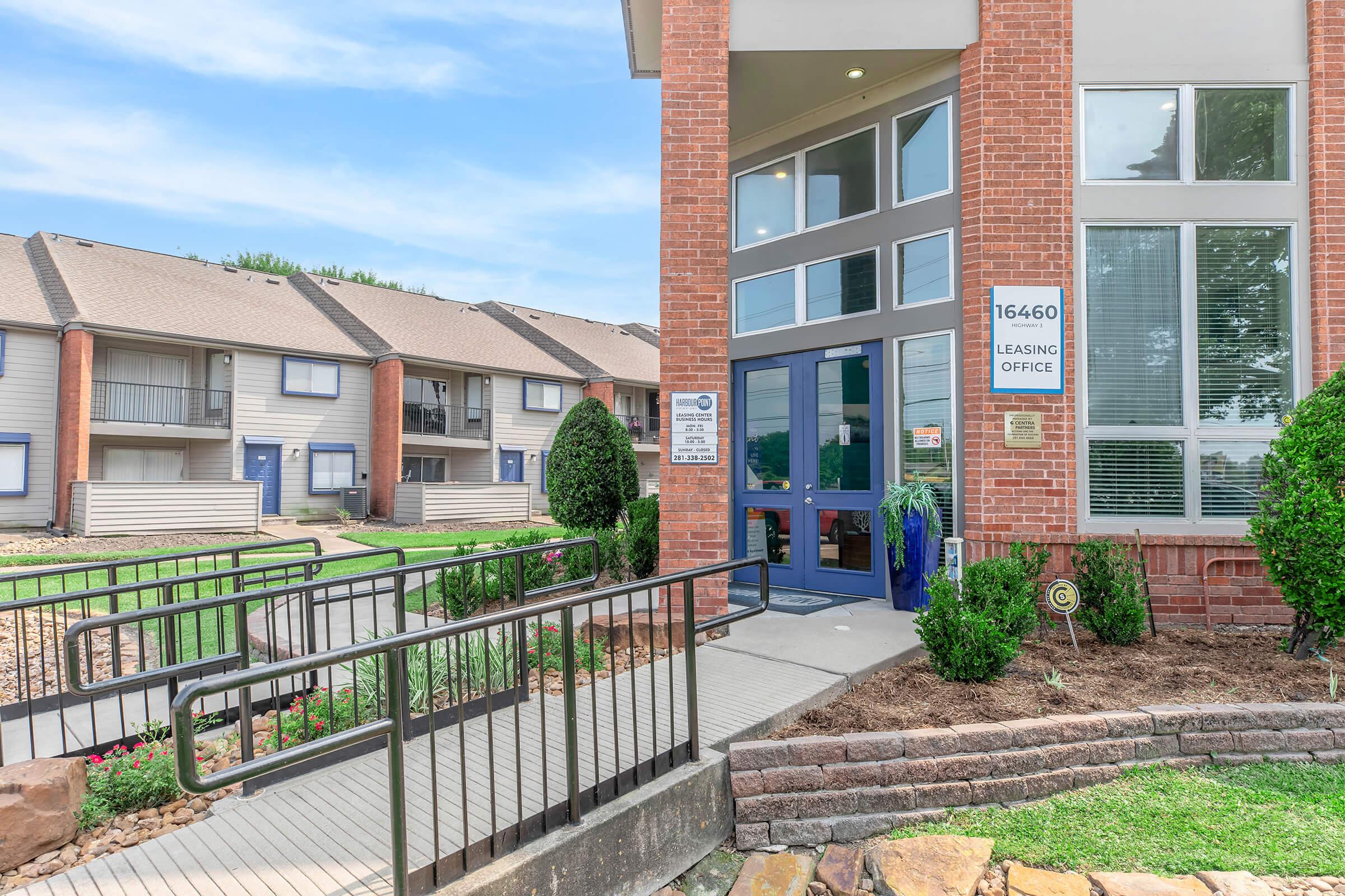 Leasing office of an apartment complex with a brick exterior, large windows, and a sign displaying the office number. There is a wheelchair-accessible ramp leading to the entrance, surrounded by neatly trimmed bushes and landscaping. In the background, additional apartment buildings are visible.