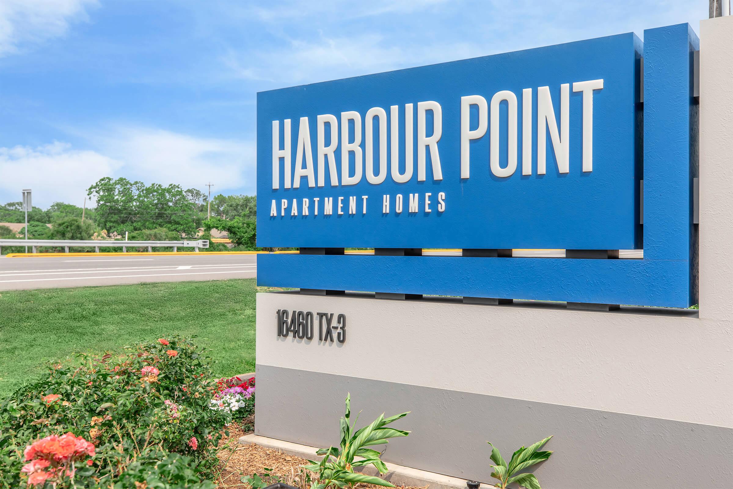 Sign for Harbour Point Apartment Homes, featuring bold blue letters on a sleek modern design. The sign is situated near a well-manicured landscape with colorful flowers and greenery in the foreground. Clear blue sky overhead adds to the inviting atmosphere. Located at 18460 TX-3.