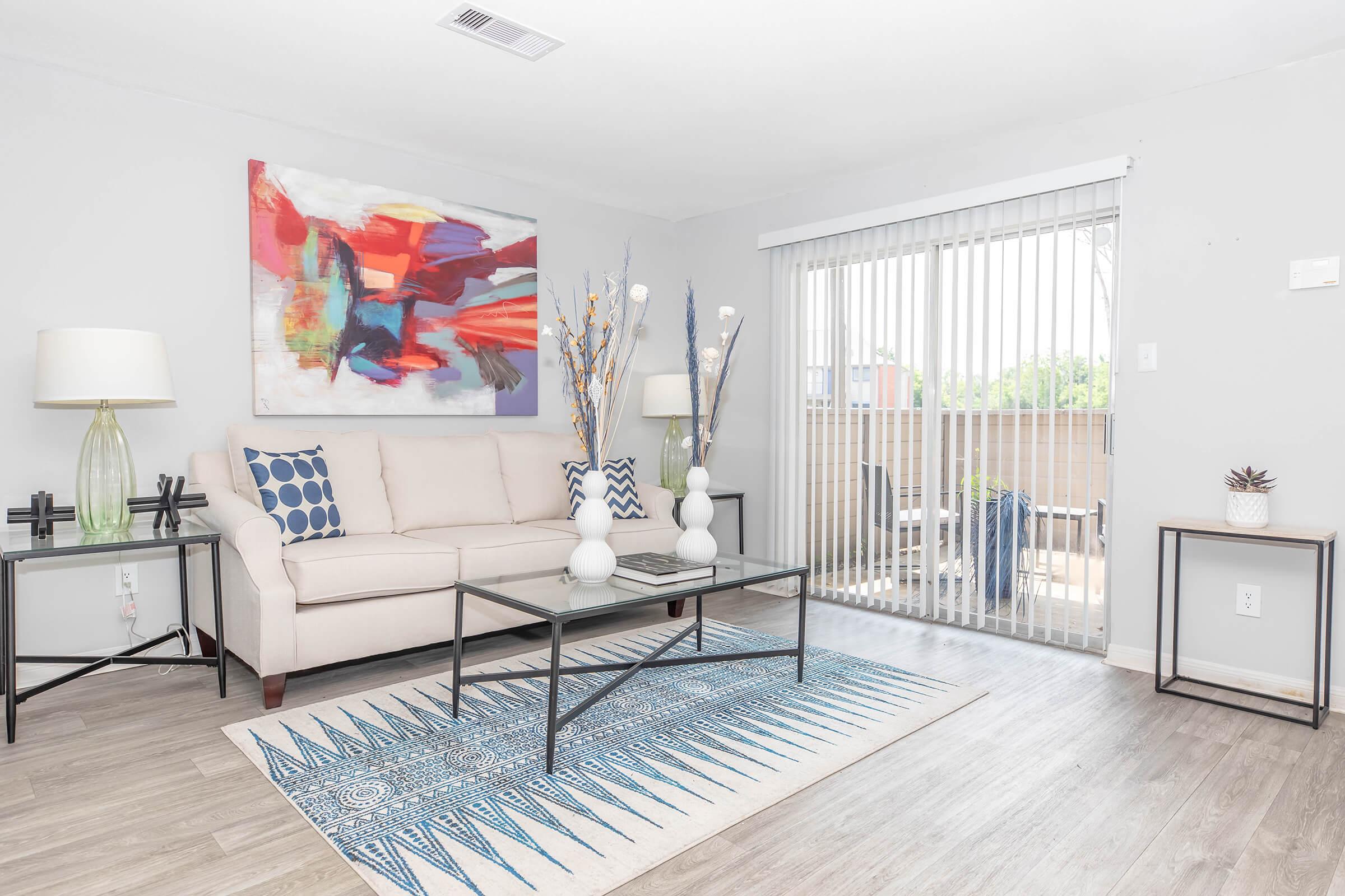A modern living room featuring a light-colored sofa, a blue and white patterned area rug, and two stylish glass-topped tables. Decorative vases with branches, colorful artwork on the wall, and vertical blinds cover a sliding glass door leading to a balcony. Neutral walls and flooring enhance the bright atmosphere.