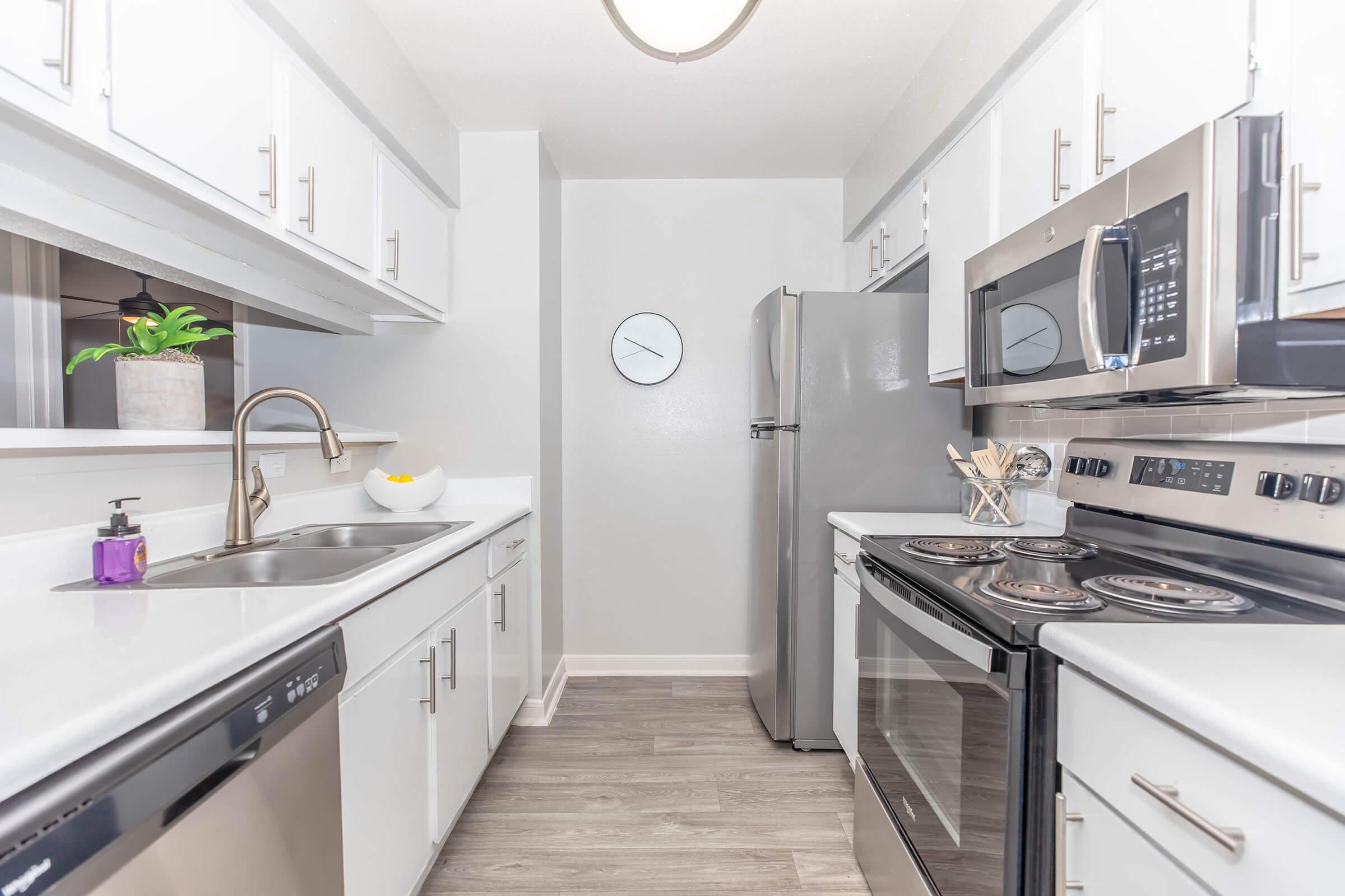 Modern kitchen with white cabinetry, stainless steel appliances including a microwave and oven, a refrigerator, and a double sink. A plant in a pot adds a touch of green, while a wall clock maintains a minimalist aesthetic. The flooring is light wood, contributing to a bright and airy feel.