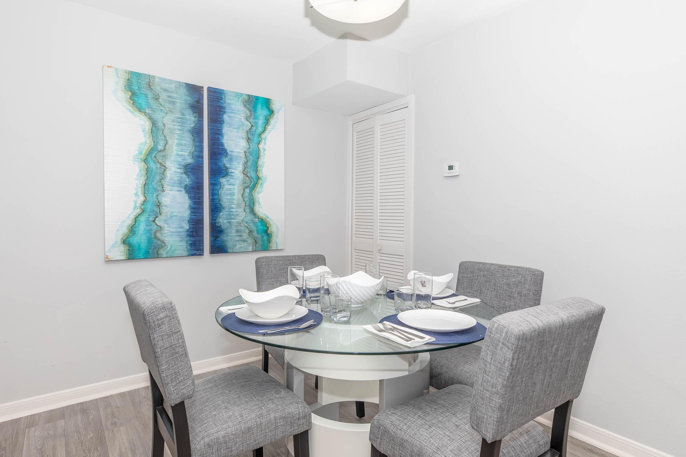 A modern dining area featuring a round glass table surrounded by four gray upholstered chairs. The table is set with white dishware and blue placemats. A large abstract artwork with blue and green hues hangs on the wall, and there's a white decorative bowl at the center of the table.