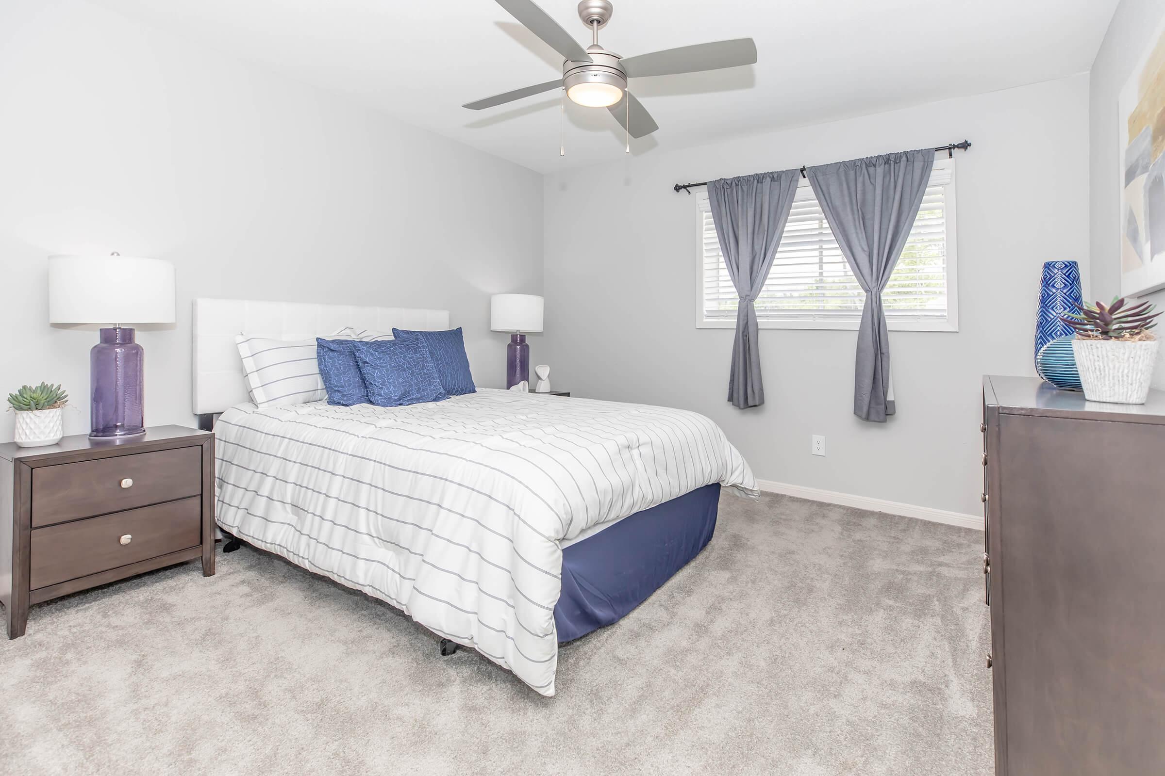 A neatly arranged bedroom featuring a bed with a white and blue striped comforter, flanked by two nightstands with lamps. There are blue pillows on the bed and decorative accents on the nightstands. The walls are light gray, with a window dressed in gray curtains, and a dark chest of drawers in the corner.
