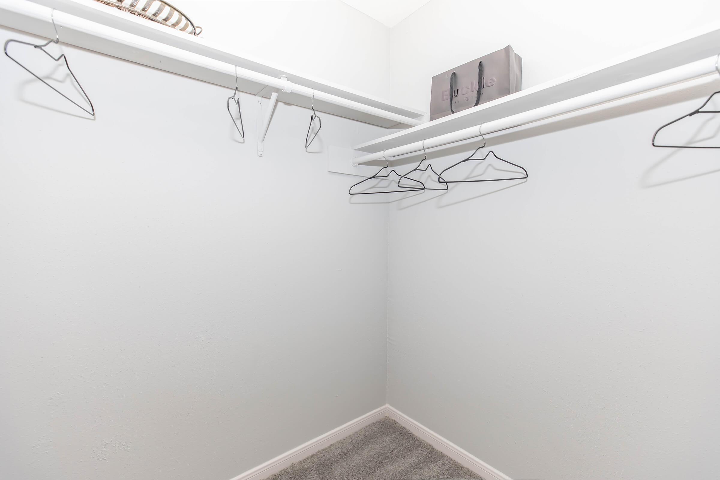 A minimalistic closet with white walls and a carpeted floor. Several black wire hangers hang from two horizontal rods, and a grey box is placed on a shelf at the back. The space appears organized and uncluttered.