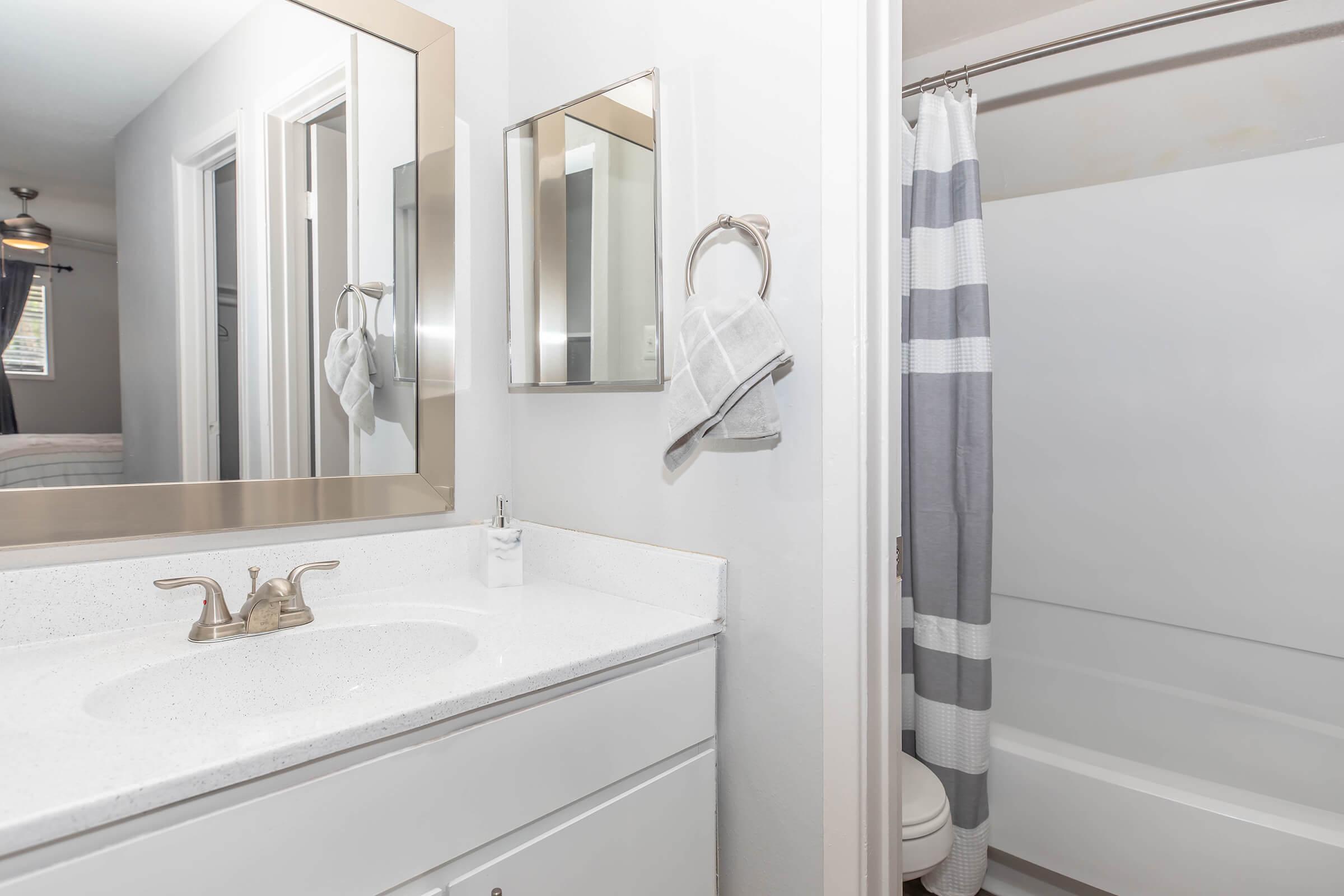 A modern bathroom featuring a white countertop with a sink, a large mirror above, and a towel rack with a gray towel. The shower area has a clear glass door and a gray-striped curtain. Light walls and a clean design create a bright and inviting space.