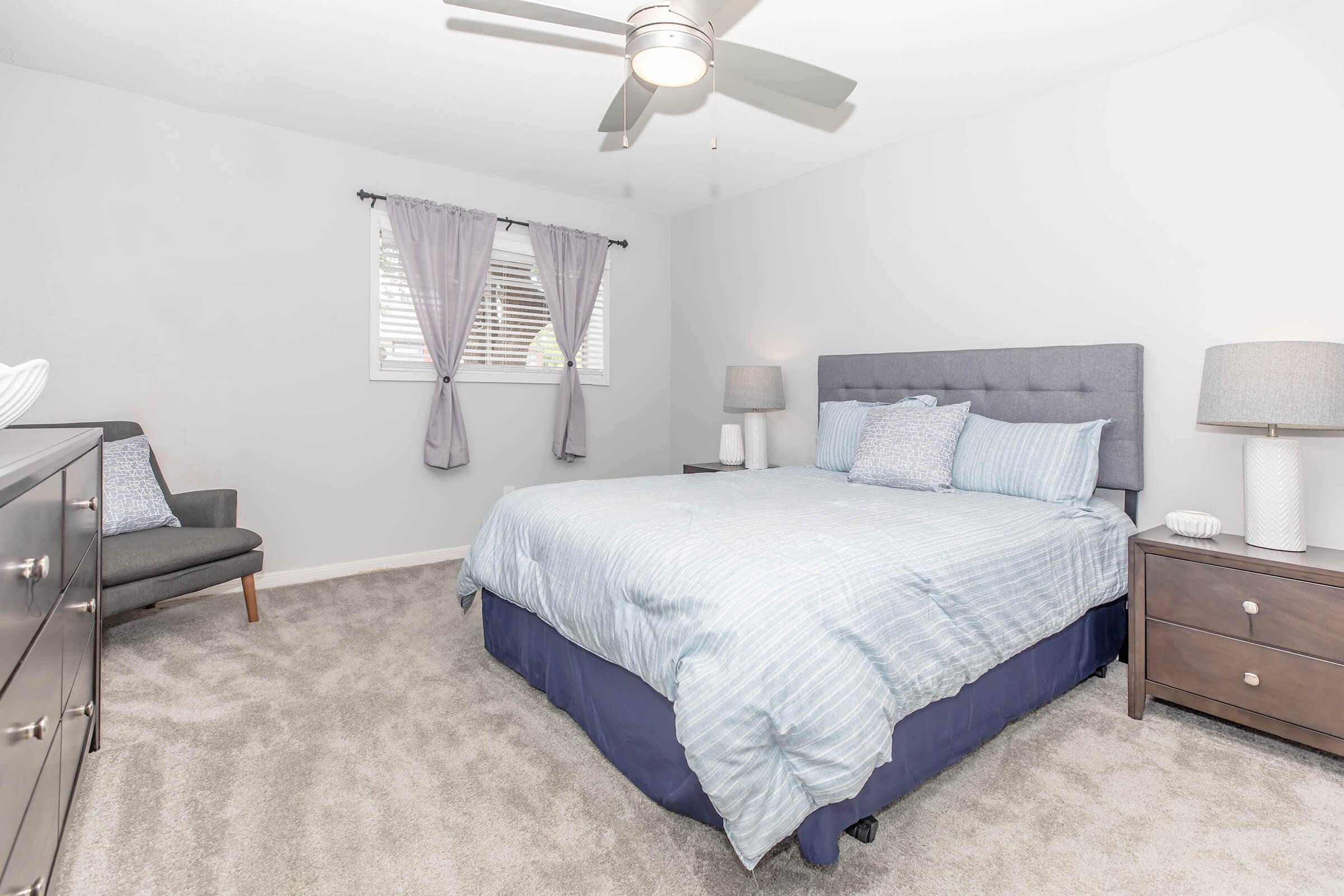 A cozy bedroom featuring a king-sized bed with light blue striped bedding, a gray upholstered headboard, and two nightstands with lamps. Natural light filters through sheer curtains on a window. A comfortable gray chair is positioned in the corner, complementing the soft gray walls and carpet.