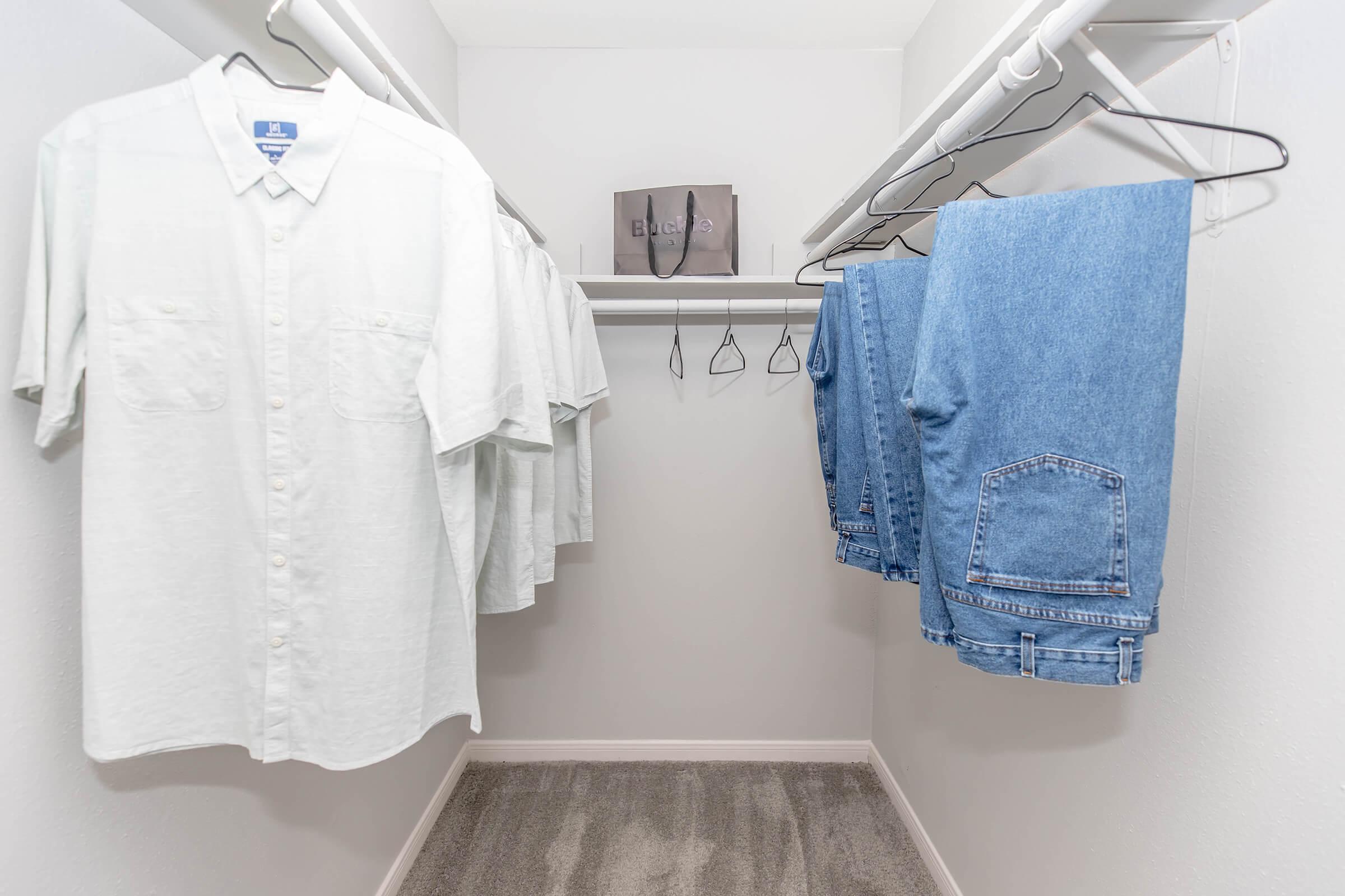 A neat closet featuring several hanging light-colored shirts and a pair of denim jeans. There is a gray handbag on a shelf above, and the closet walls and floor are painted in neutral colors, creating a tidy and organized appearance.