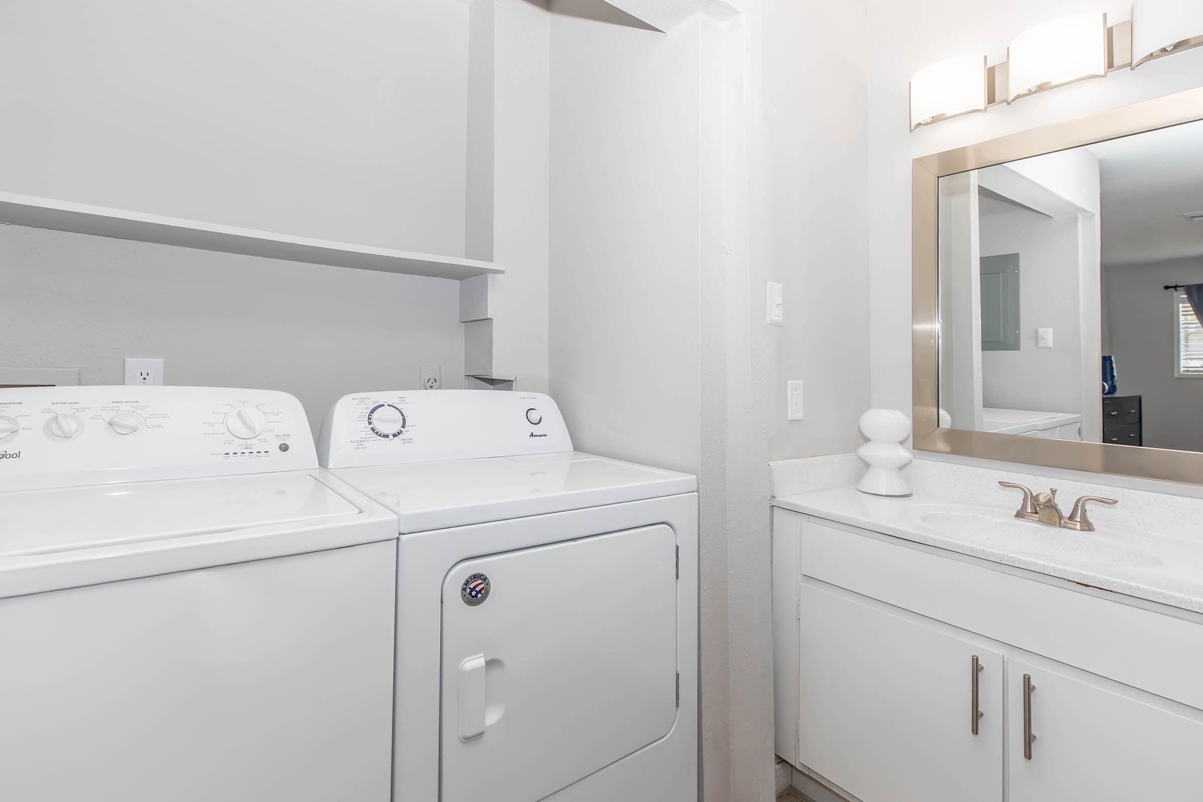 A clean laundry room featuring a stacked washer and dryer on the left, with a countertop space above. On the right, there's a bathroom vanity with a large mirror, a modern faucet, and a decorative white lamp. The walls are painted in a light color, enhancing the spacious feel of the room.