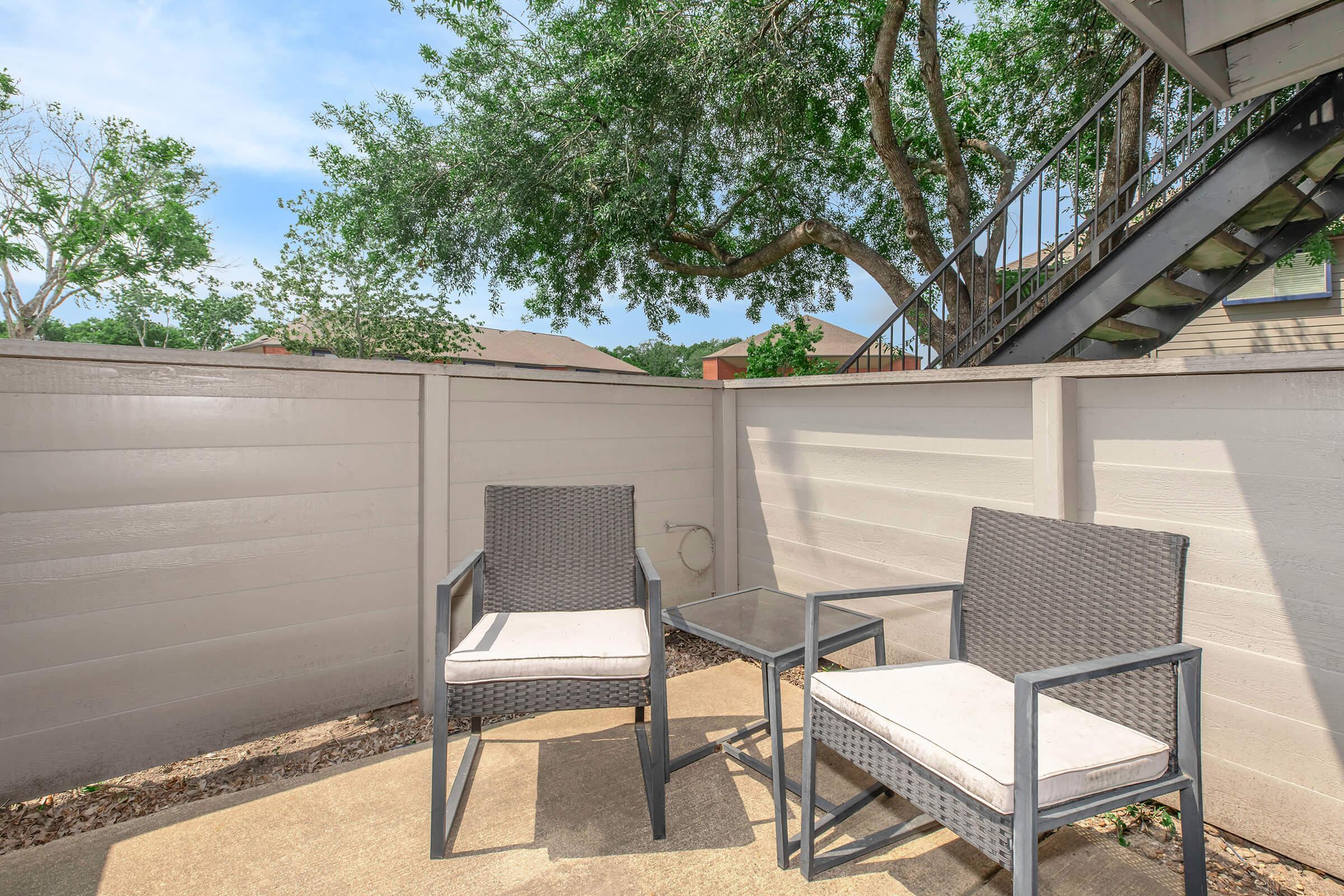 A small outdoor patio area featuring two wicker chairs with cushions and a glass side table. The space is enclosed by a low wall, with trees visible in the background and stairs leading up from one side. Bright blue sky enhances the inviting atmosphere of the setting.