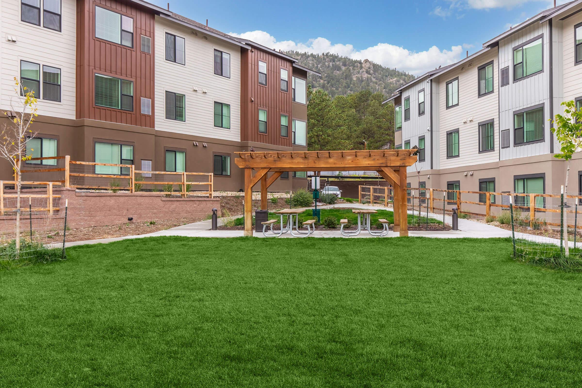 A landscaped courtyard featuring a lush green lawn, a wooden pergola with seating, and two multi-story residential buildings in the background. The setting is surrounded by trees and mountains, providing a serene outdoor space for residents. Bright blue sky and scattered clouds enhance the scenery.