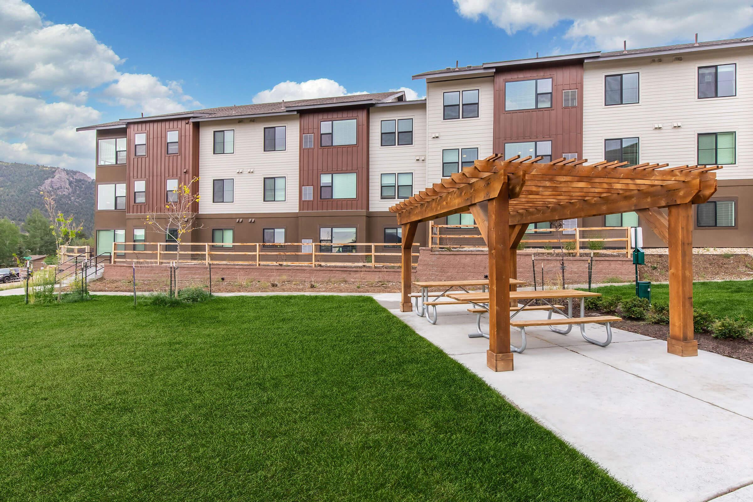 A grassy area with a wooden pergola and picnic tables in front of a modern multi-story building. The building features a mix of light and dark siding with multiple windows, set against a backdrop of blue sky and clouds.
