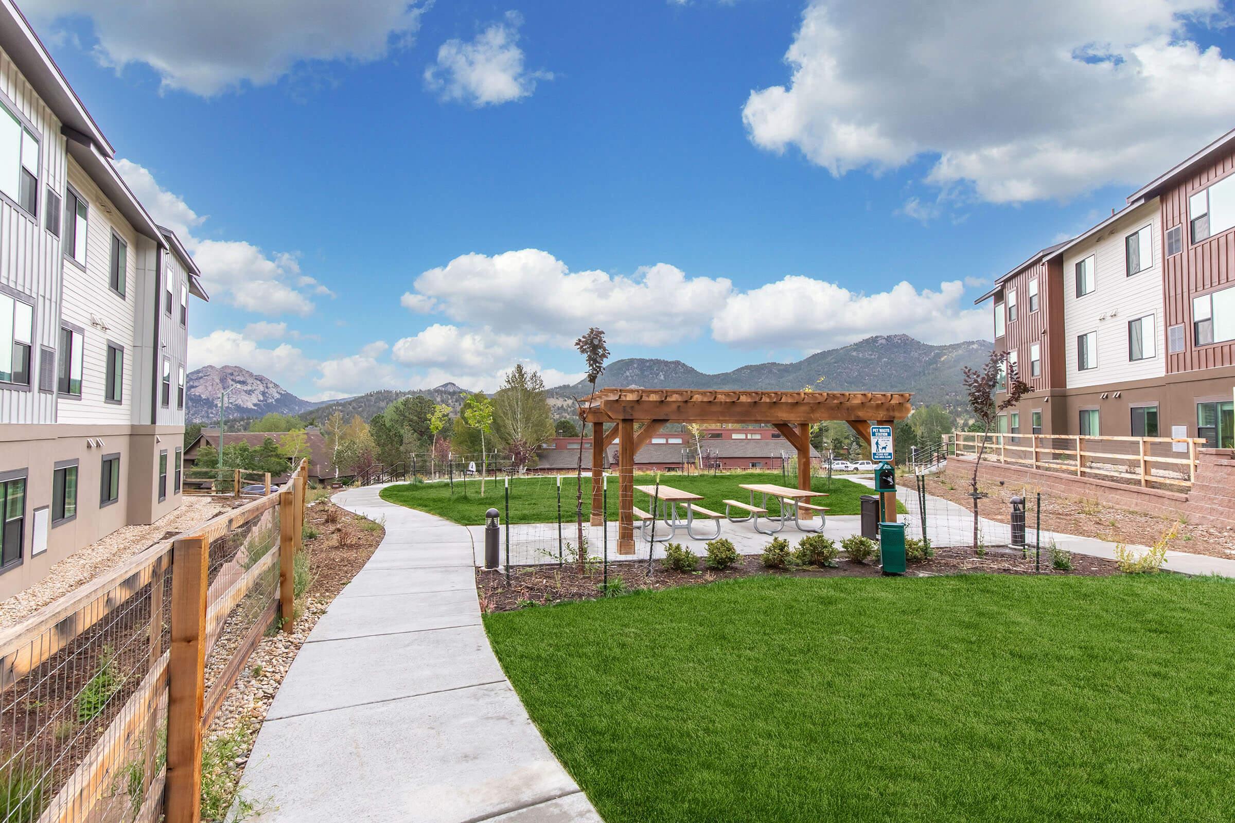 A landscaped pathway winds through a green lawn between two modern buildings. A wooden pergola stands in the open area, surrounded by picnic tables. Mountains are visible in the background under a bright blue sky with fluffy clouds. The scene conveys a serene and inviting outdoor space.