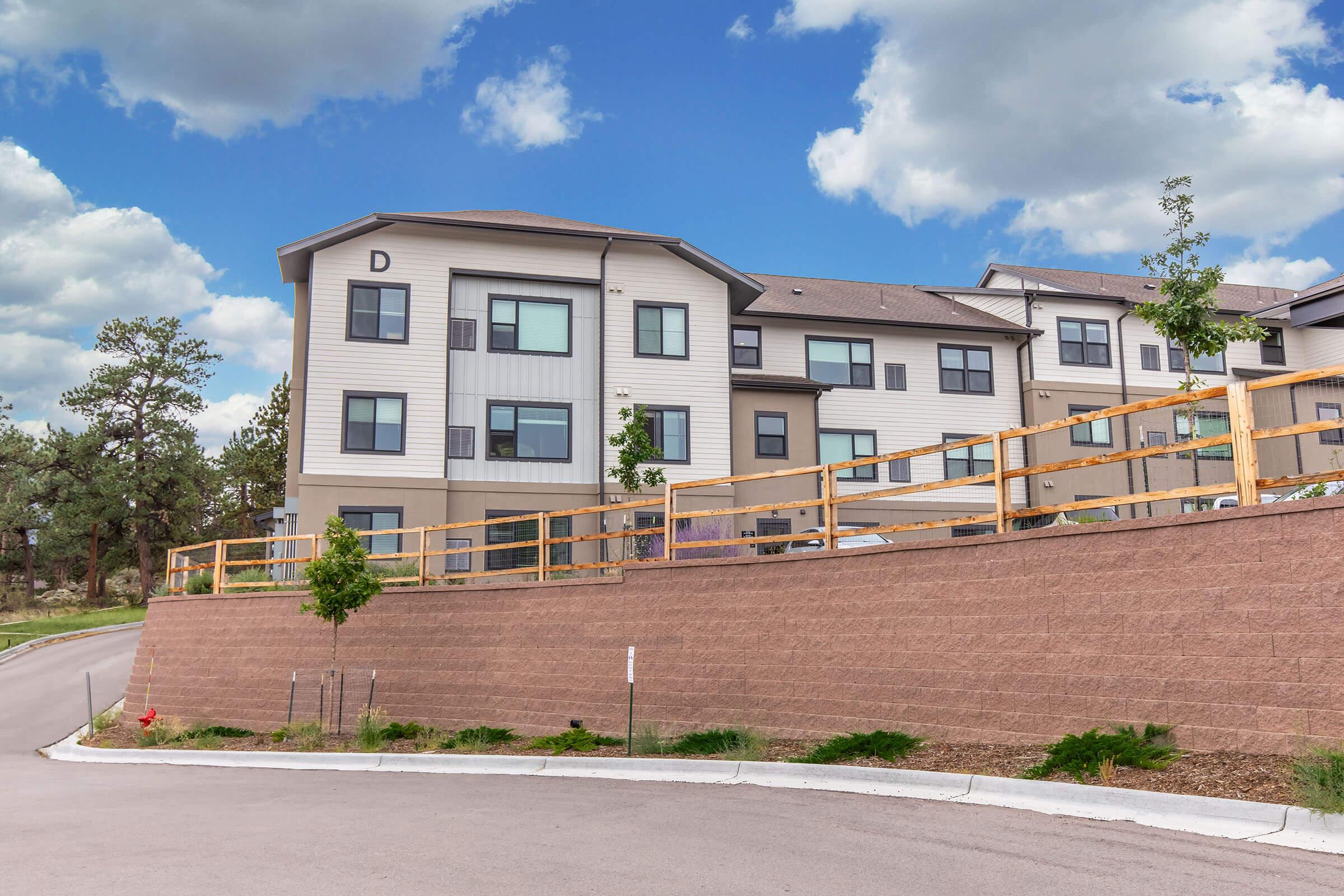 Modern multi-unit residential building with a light-colored exterior, situated on a landscaped sloped lot. Features a wooden fence along the front, surrounded by greenery, under a blue sky with scattered clouds. A gently curving road leads up to the entrance.