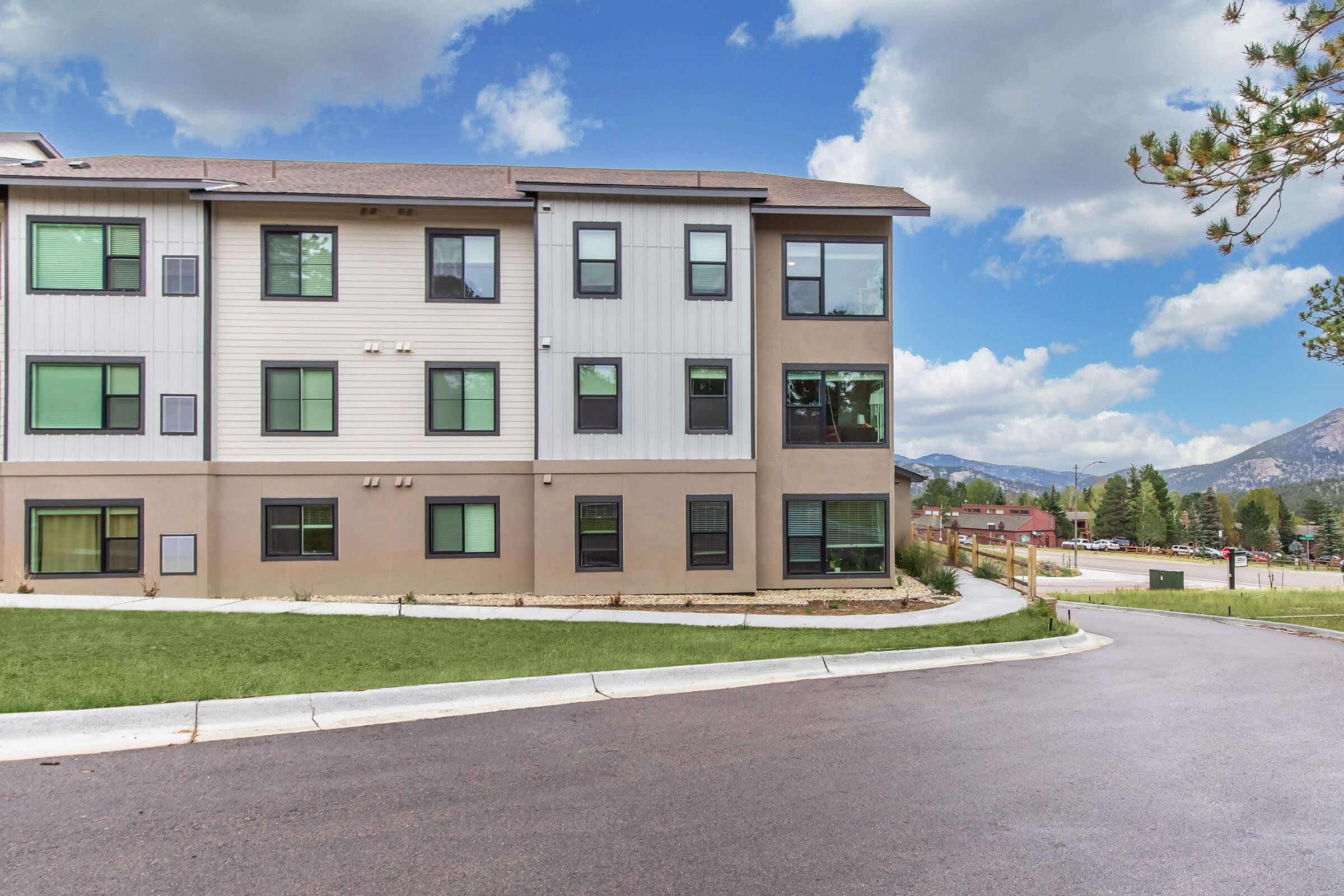 A modern multi-story building with a mix of light and dark exterior siding. The structure features multiple windows, a landscaped area in front, and a paved road leading up to it. In the background, there are mountains and a blue sky with clouds, indicating a serene environment.