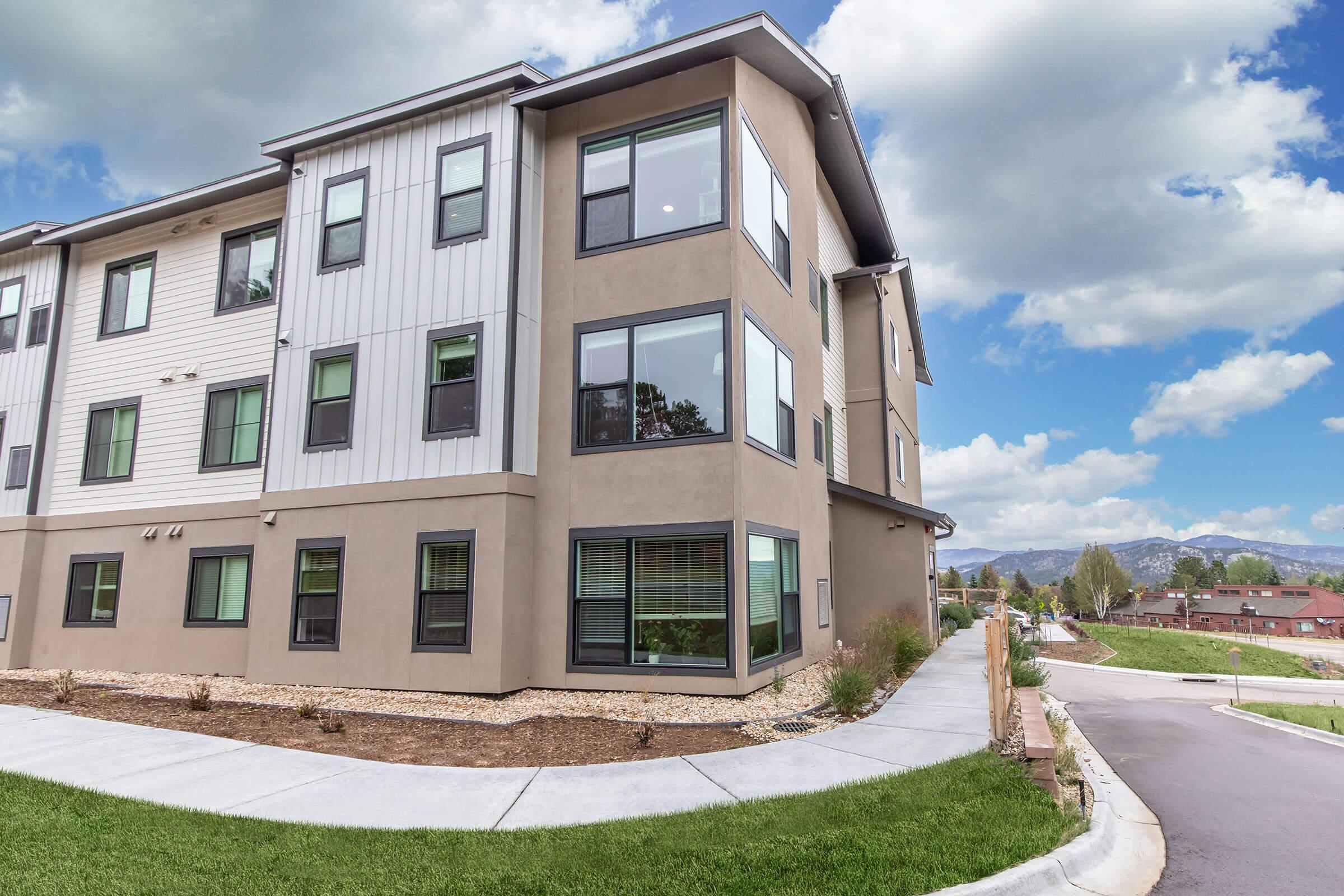 A modern multi-story residential building with a mix of beige and gray exterior walls, large windows, and a landscaped area featuring grass and small plants. The building is situated on a slight elevation with a walkway leading from the street, under a partly cloudy sky with distant mountains visible.