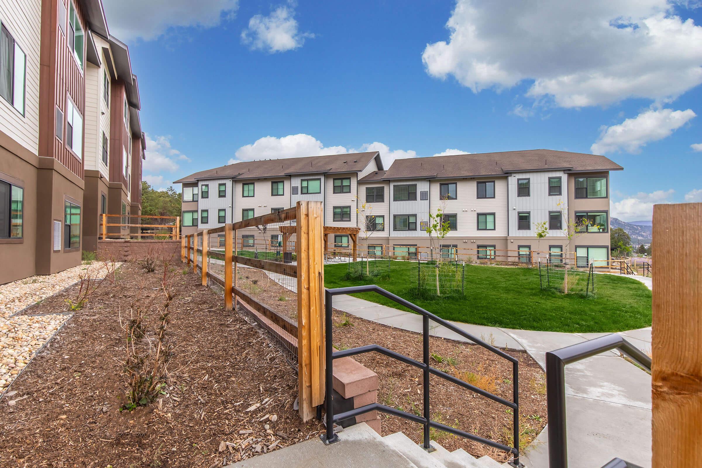 View of a multi-story apartment complex with a well-maintained exterior. In the foreground, there is a paved path leading down a set of stairs with wooden railings. Grass and small trees are visible in the landscaped area, set against a backdrop of blue sky with fluffy clouds.
