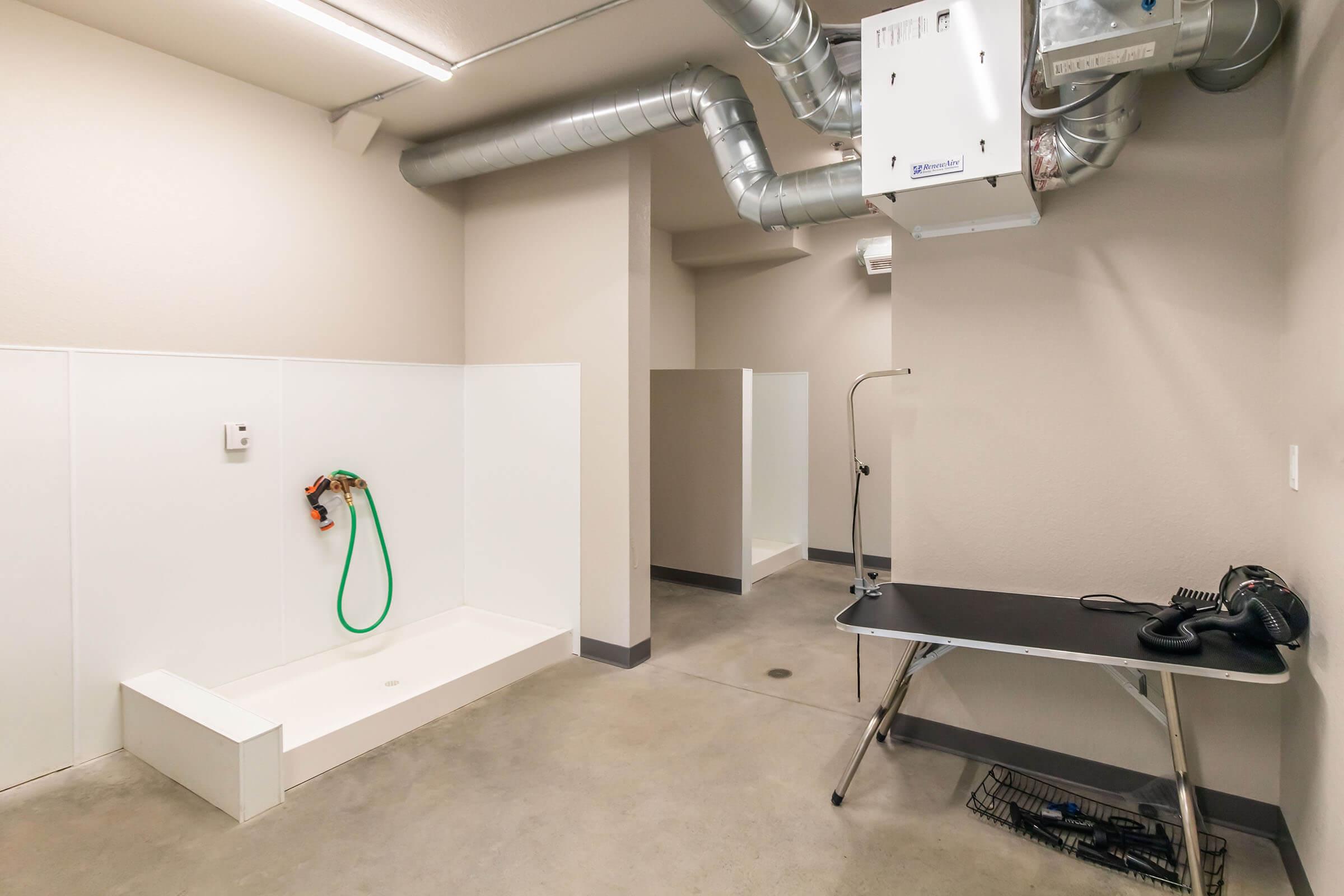 A spacious indoor area featuring a shower installation with a green hose, gray concrete flooring, and ventilation ducts on the ceiling. There are two partitions likely for changing or privacy, and a metal table with a phone and a black basket nearby. The walls are painted light gray.