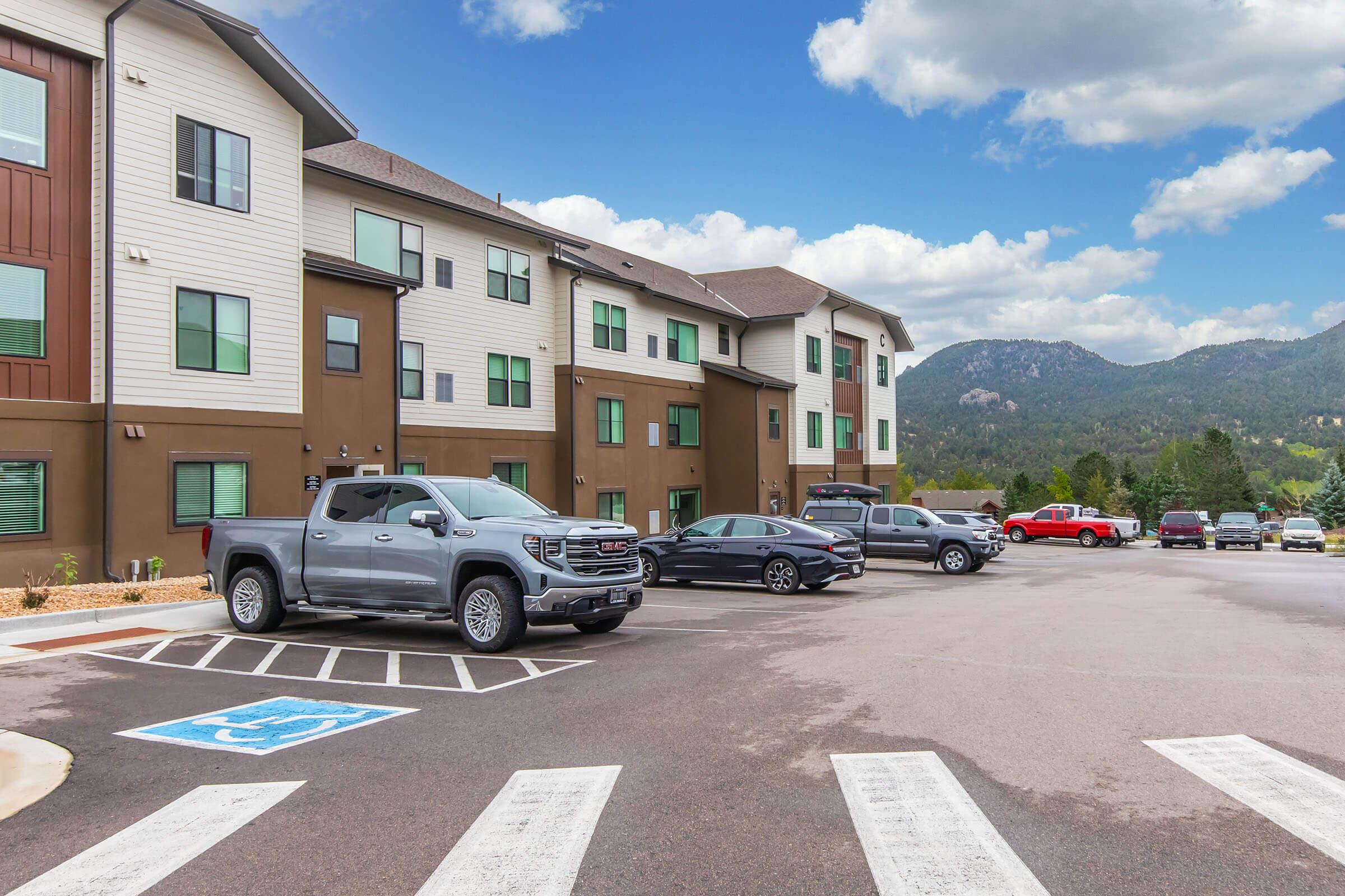 A parking lot in front of a multi-story hotel or apartment building, featuring several parked vehicles, including a silver pickup truck and a red truck. The backdrop includes green trees and mountains under a partly cloudy sky.