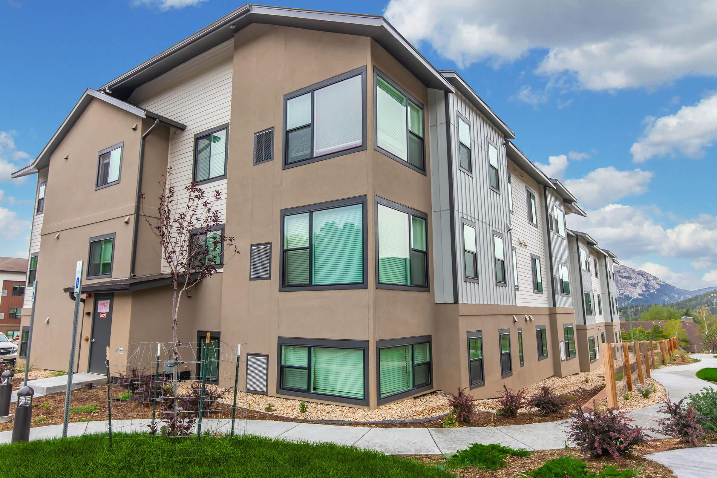 A modern three-story residential building with large windows, surrounded by green landscaping and trees. The exterior features a combination of light and dark earthy tones. The sky is partly cloudy, and the area appears well-maintained and inviting.