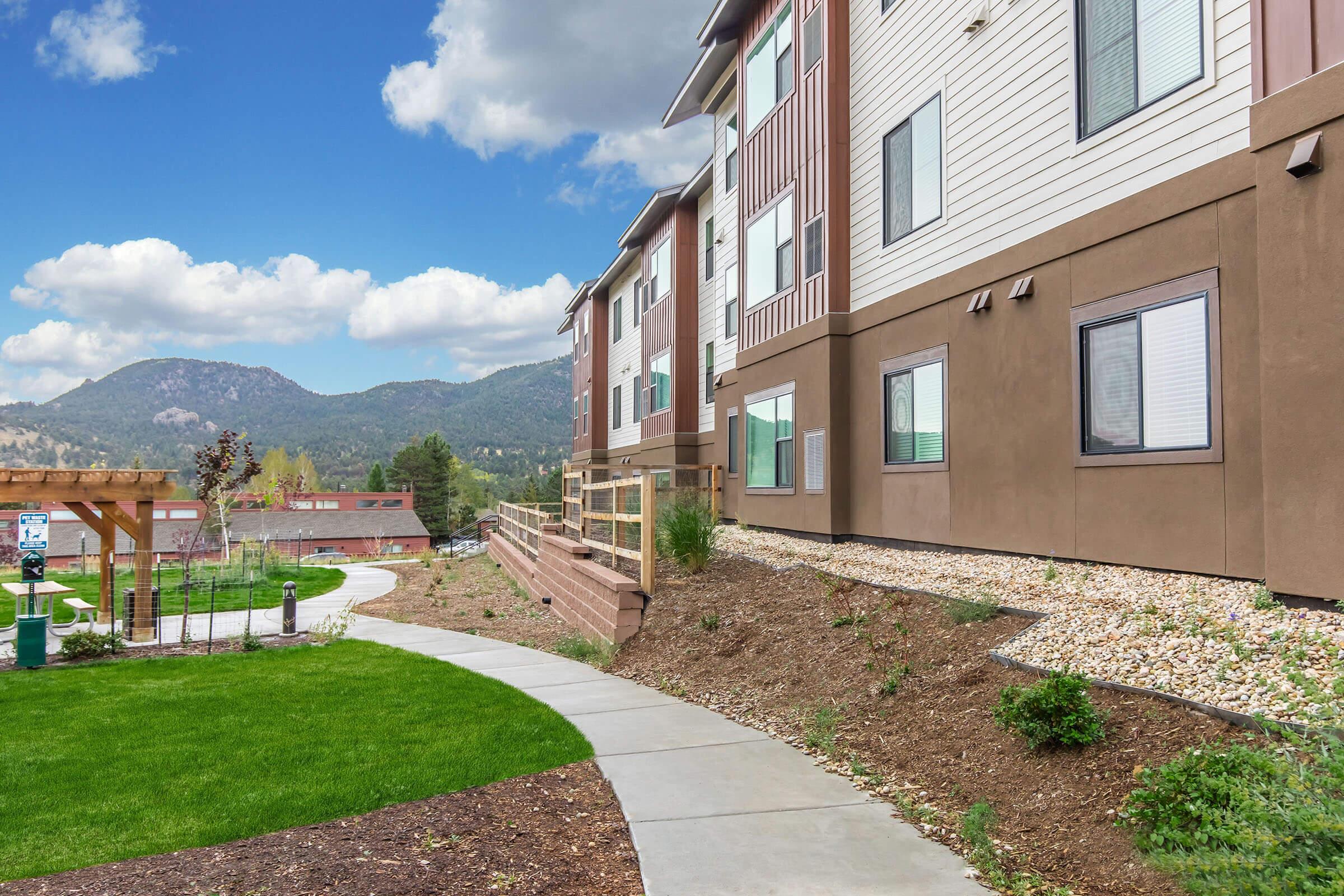A modern apartment complex set against a mountainous backdrop. The image features a well-maintained landscape with a walkway leading to the building, green grass, and a wooden pergola in the background, under a partly cloudy blue sky. The setting conveys a blend of nature and contemporary living.