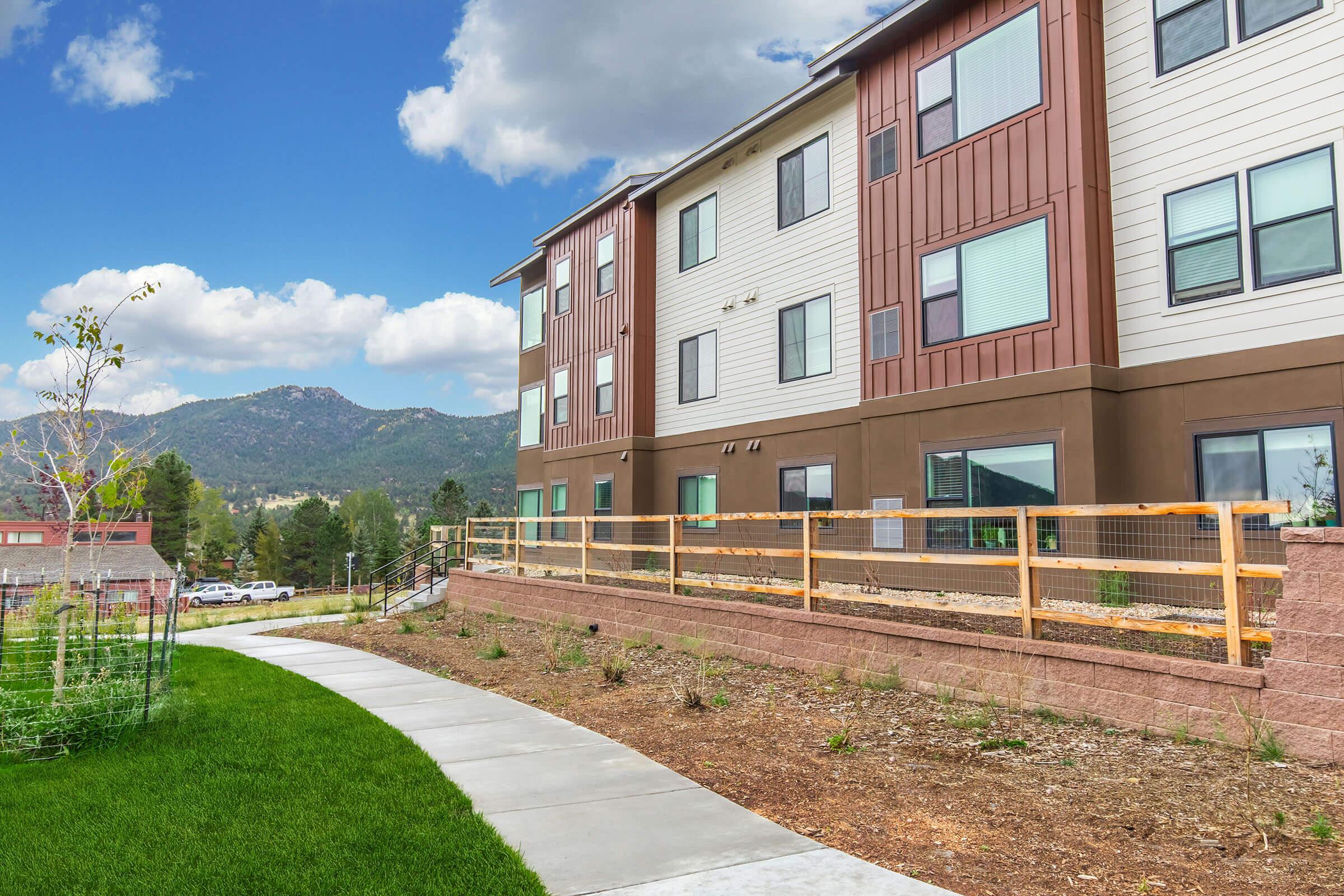 A view of a modern multi-story apartment building with a light brown and beige exterior, situated in a scenic area. The building is surrounded by a landscaped yard with grass and a walking path, set against a backdrop of mountains and a partly cloudy sky.