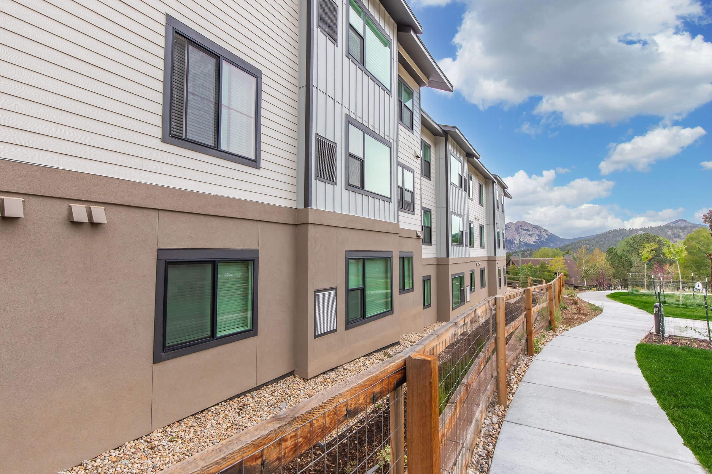 View of a modern apartment building featuring multiple levels with a combination of beige and gray siding. A paved walkway curves alongside the building, bordered by wooden fencing and landscaped areas with greenery and rocks. In the background, there are mountains and partly cloudy skies.