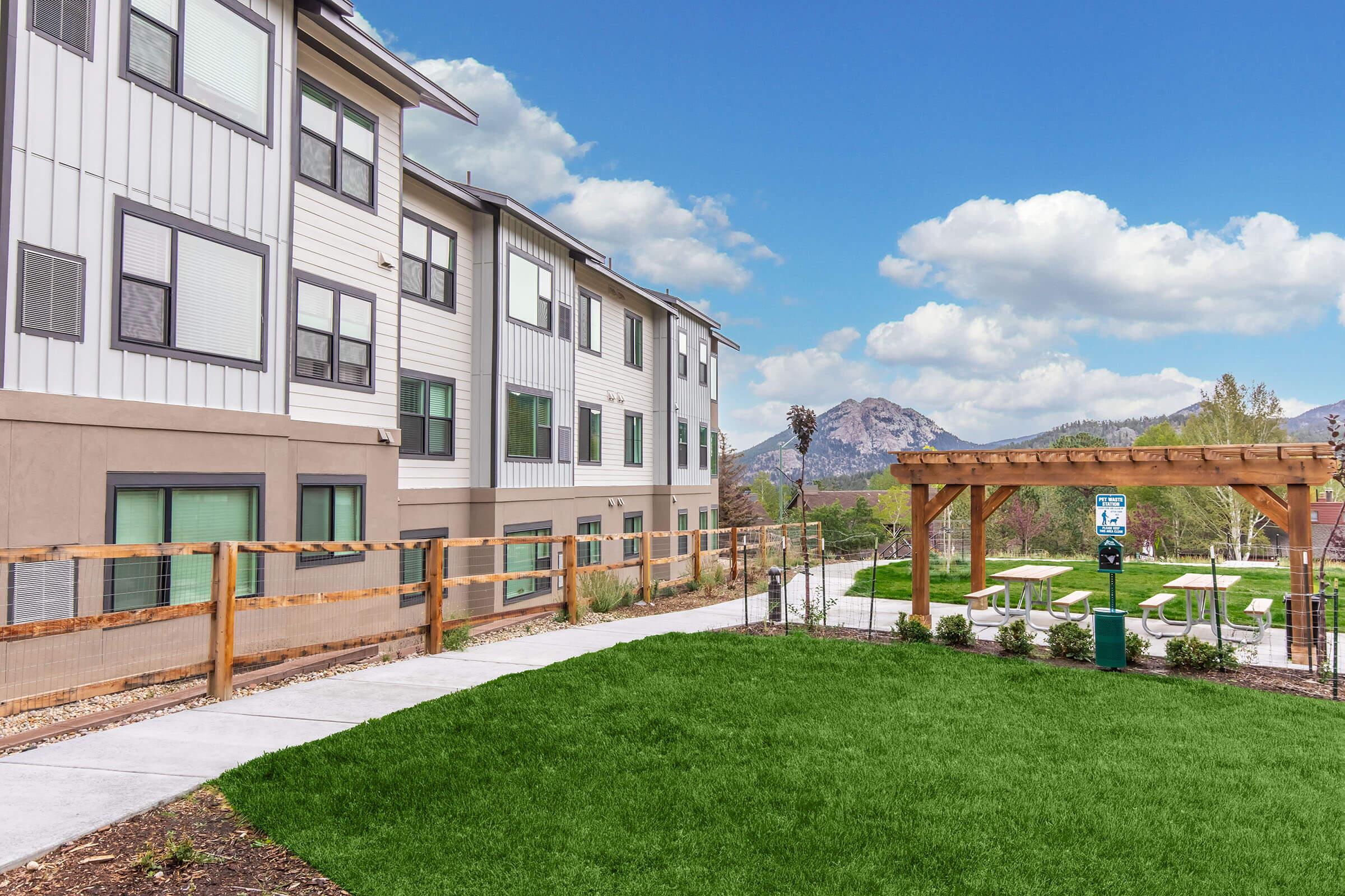 A modern residential complex with multiple stories, featuring a landscaped area with green grass and picnic tables under a wooden pergola. In the background, mountains and a partly cloudy sky can be seen, creating a scenic backdrop for the property.