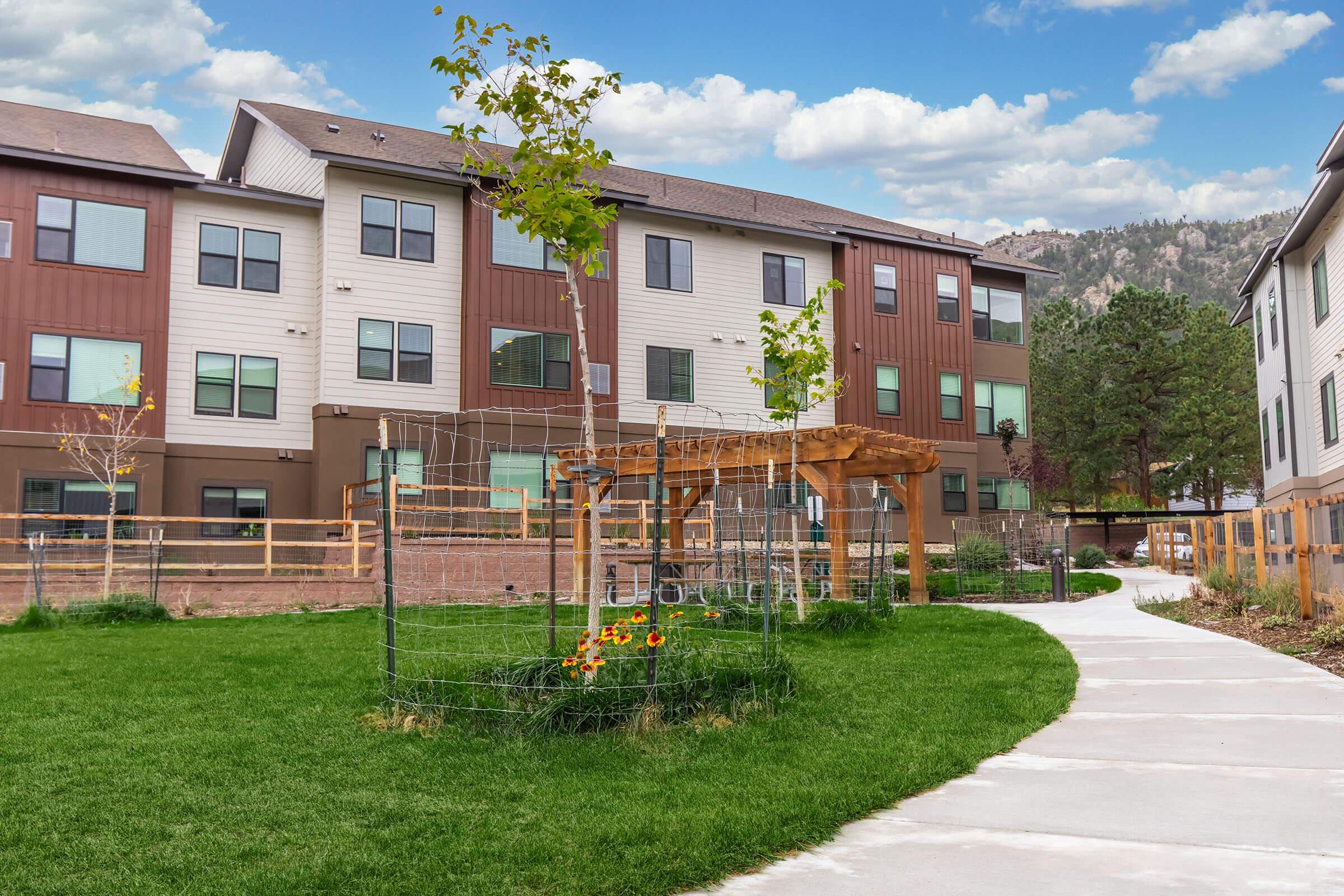 A landscaped courtyard featuring freshly planted trees, a wooden pergola, and a garden area with sunflowers. A winding pathway made of concrete leads through the green grass, with modern apartment buildings visible in the background against a partly cloudy sky.