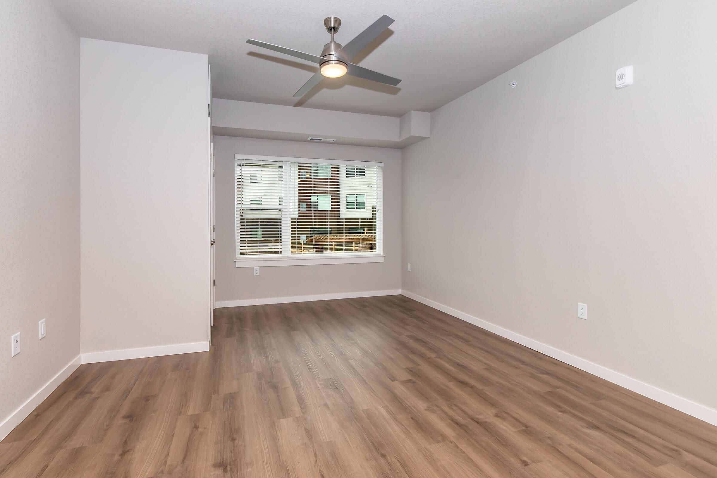 A spacious, empty room with light-colored walls and a wooden floor. A ceiling fan is mounted in the center. Natural light streams through a large window with blinds, offering a view of the outdoors. The room has a minimalistic design, making it suitable for various purposes.