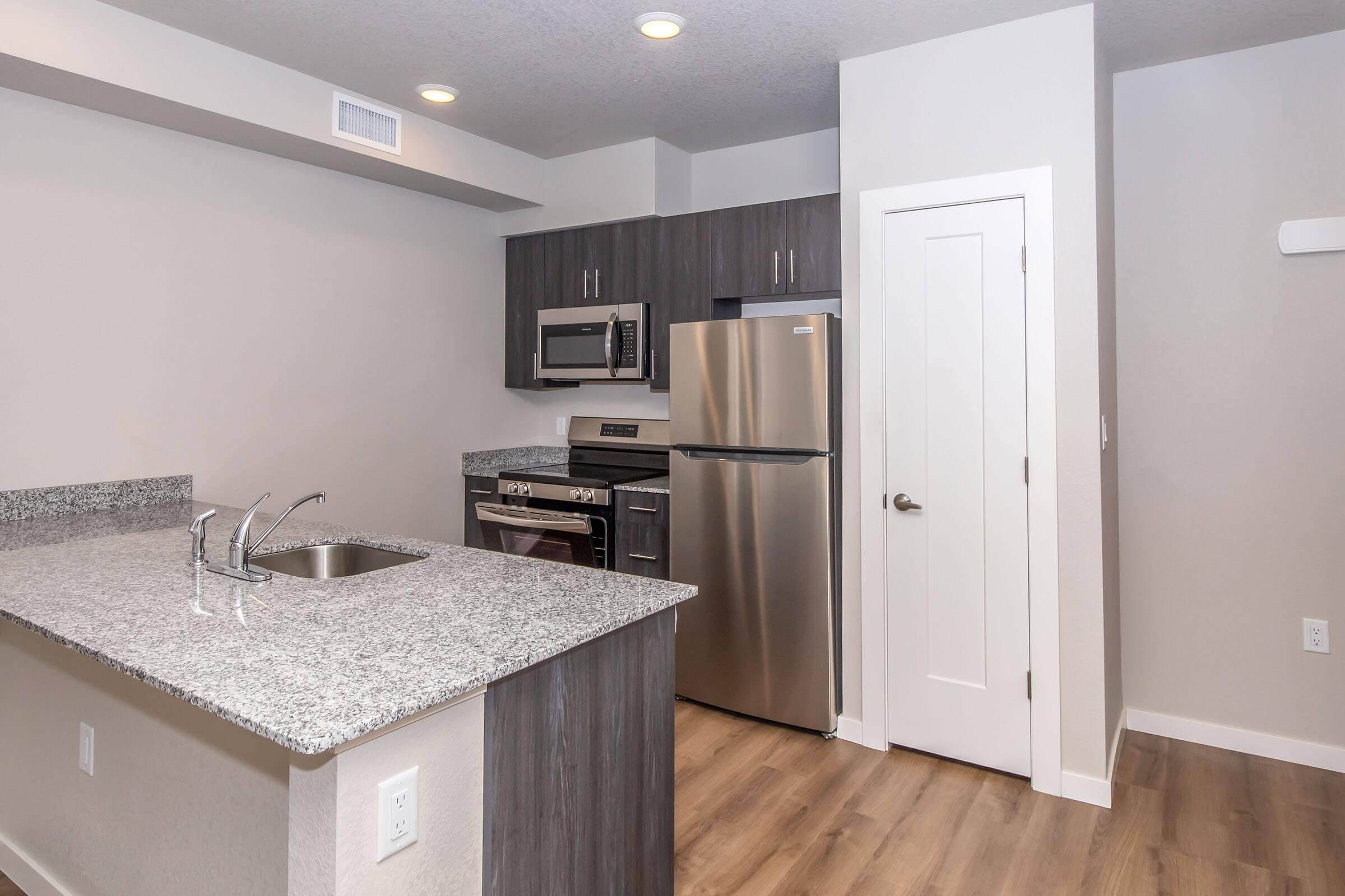 Contemporary kitchen with stainless steel appliances, including a microwave and refrigerator, set against dark wood cabinetry. The countertop is a light-colored granite, and the floor is a warm-tone laminate. The room features recessed lighting and is painted in neutral tones, creating a modern and inviting atmosphere.