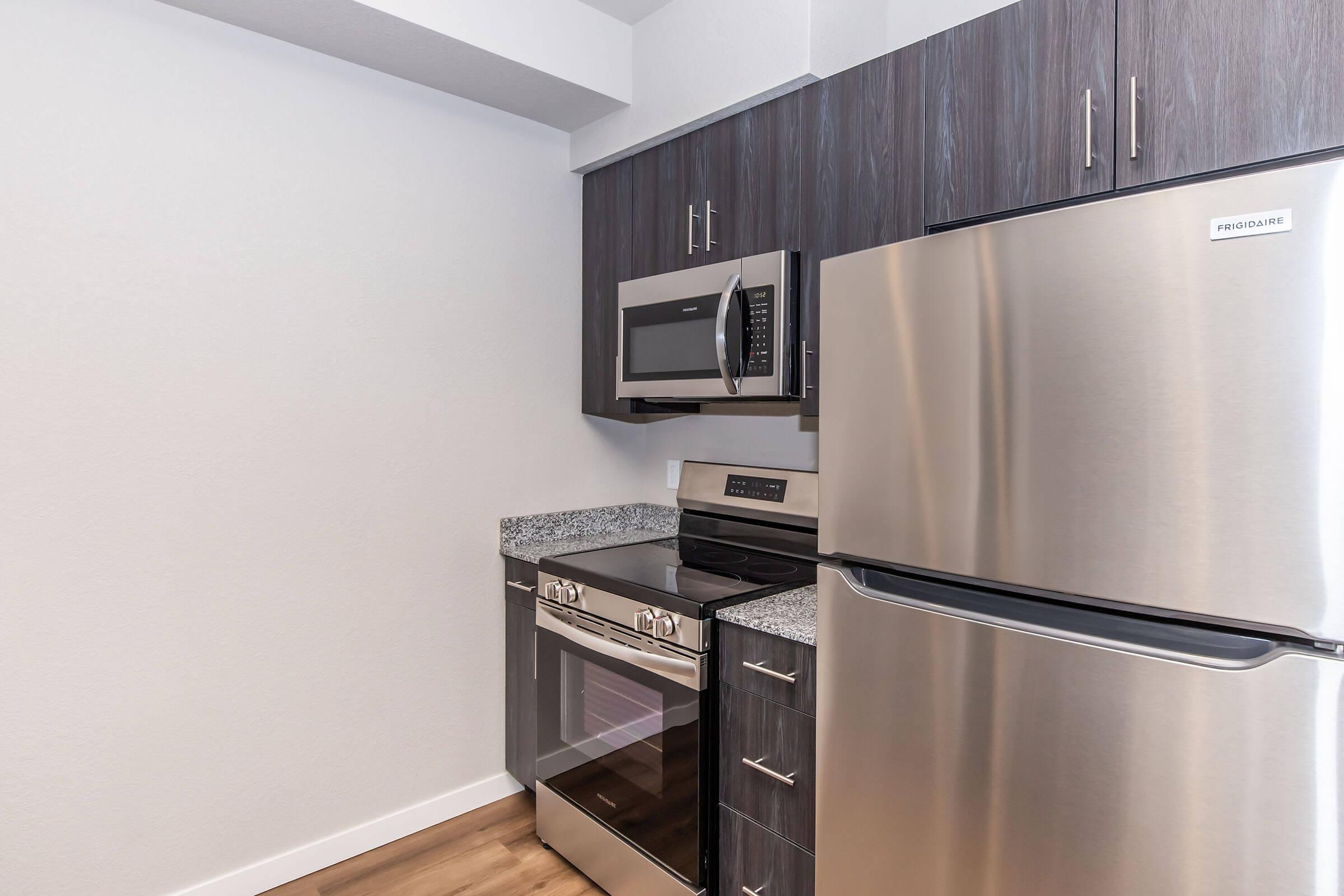Modern kitchen featuring stainless steel appliances, including a microwave, oven, and refrigerator. The cabinetry is dark wood with sleek handles, and the countertop is a light granite. The floor is a warm wood finish, creating a contemporary and inviting space.